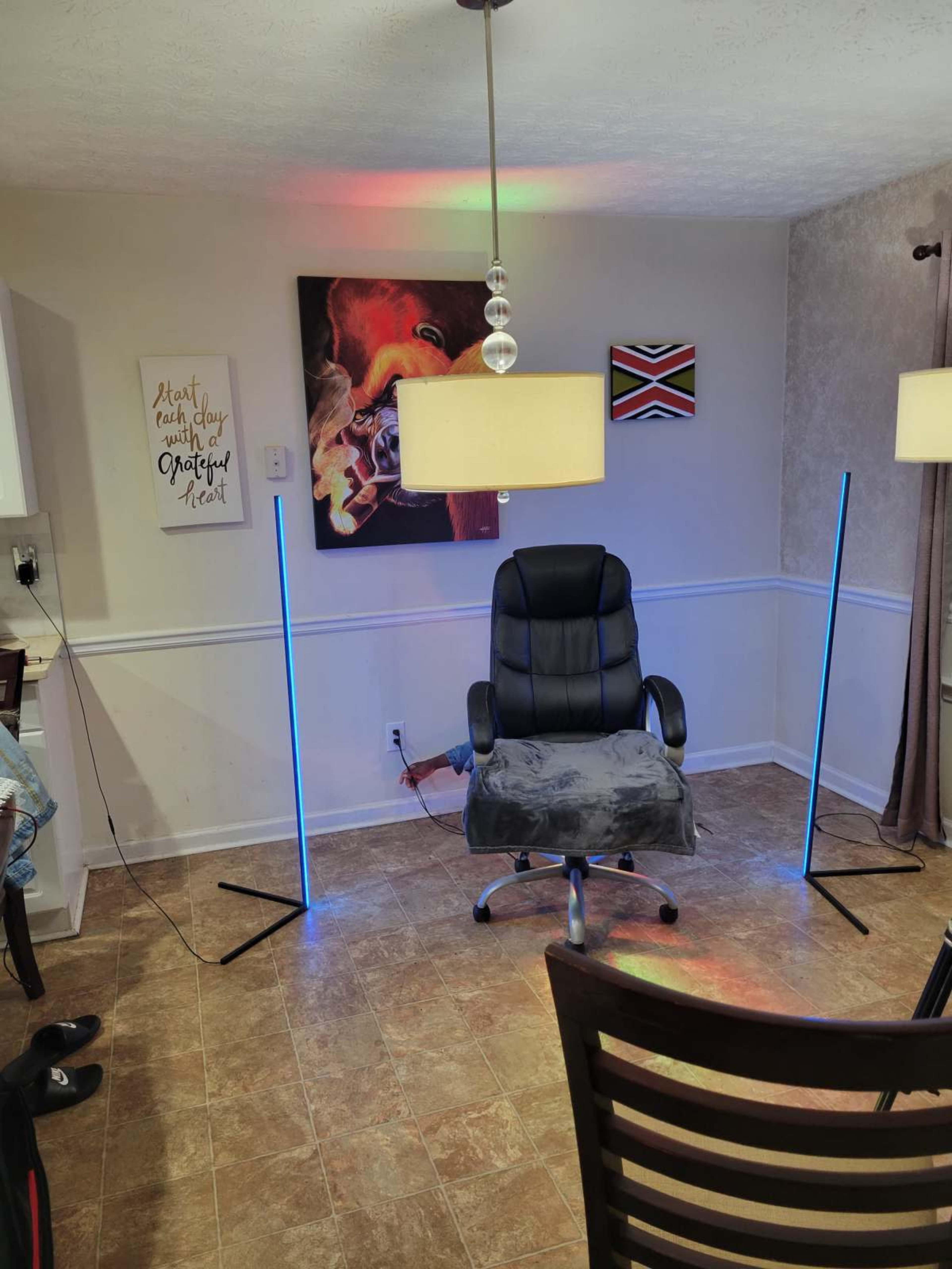 The image shows a well-lit room with a black office chair positioned between two upright light stands, surrounded by colorful wall art and light fixtures.