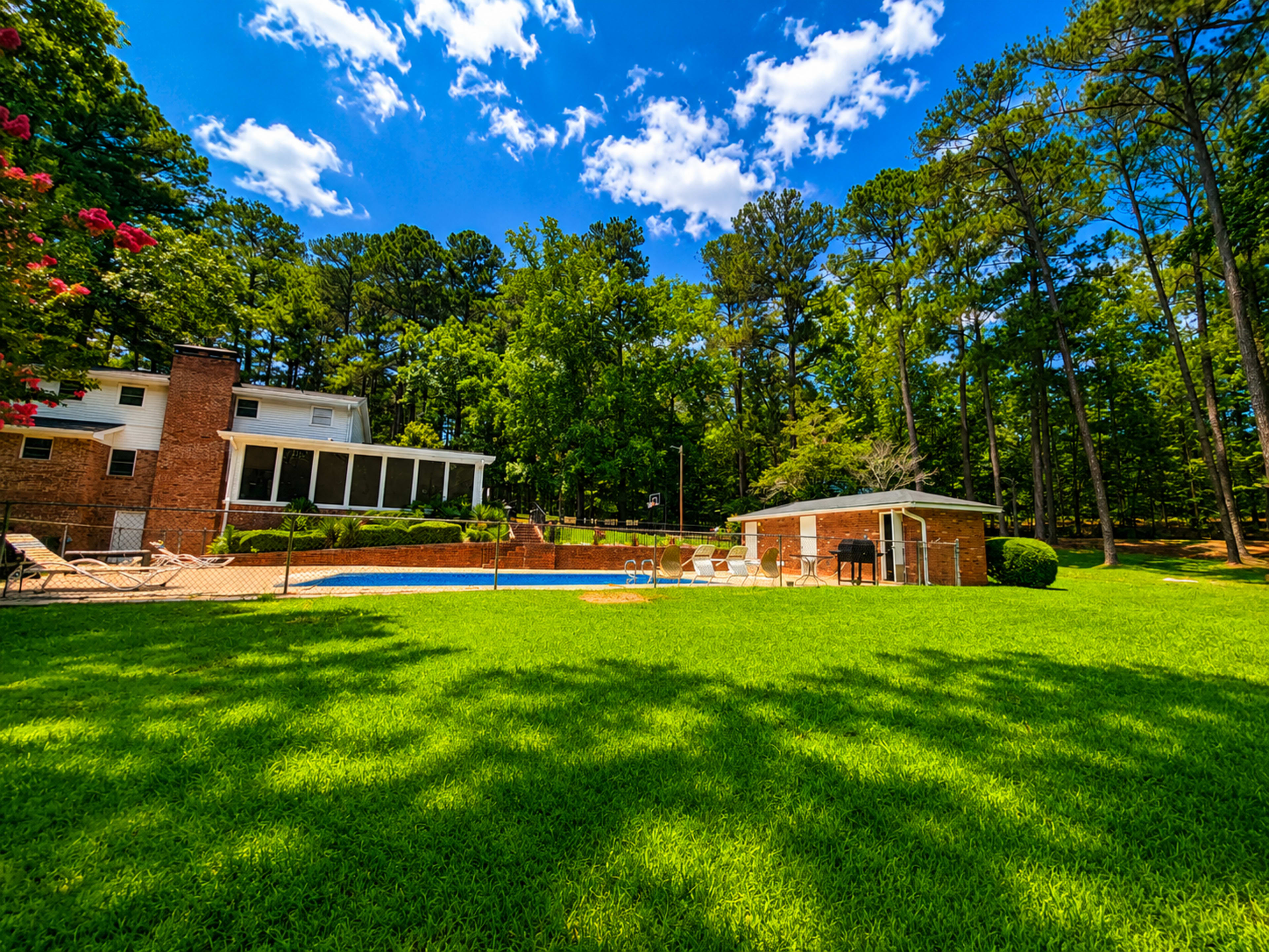 The image shows a backyard featuring a swimming pool surrounded by a lawn and trees, with a house visible in the background.