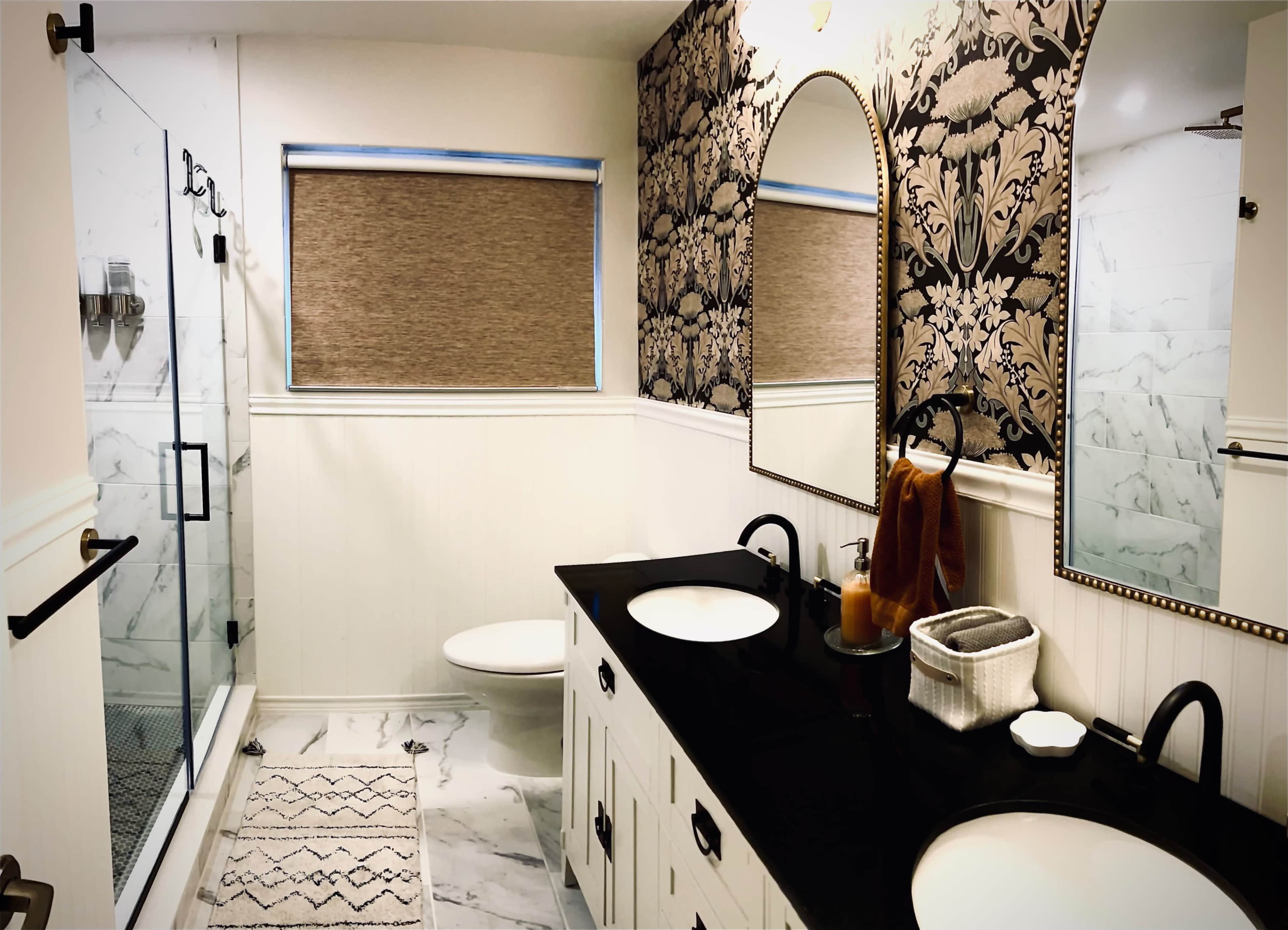 A modern bathroom with a double vanity, black countertops, two mirrors, and a glass shower enclosure, featuring floral wallpaper and a window with a shade.