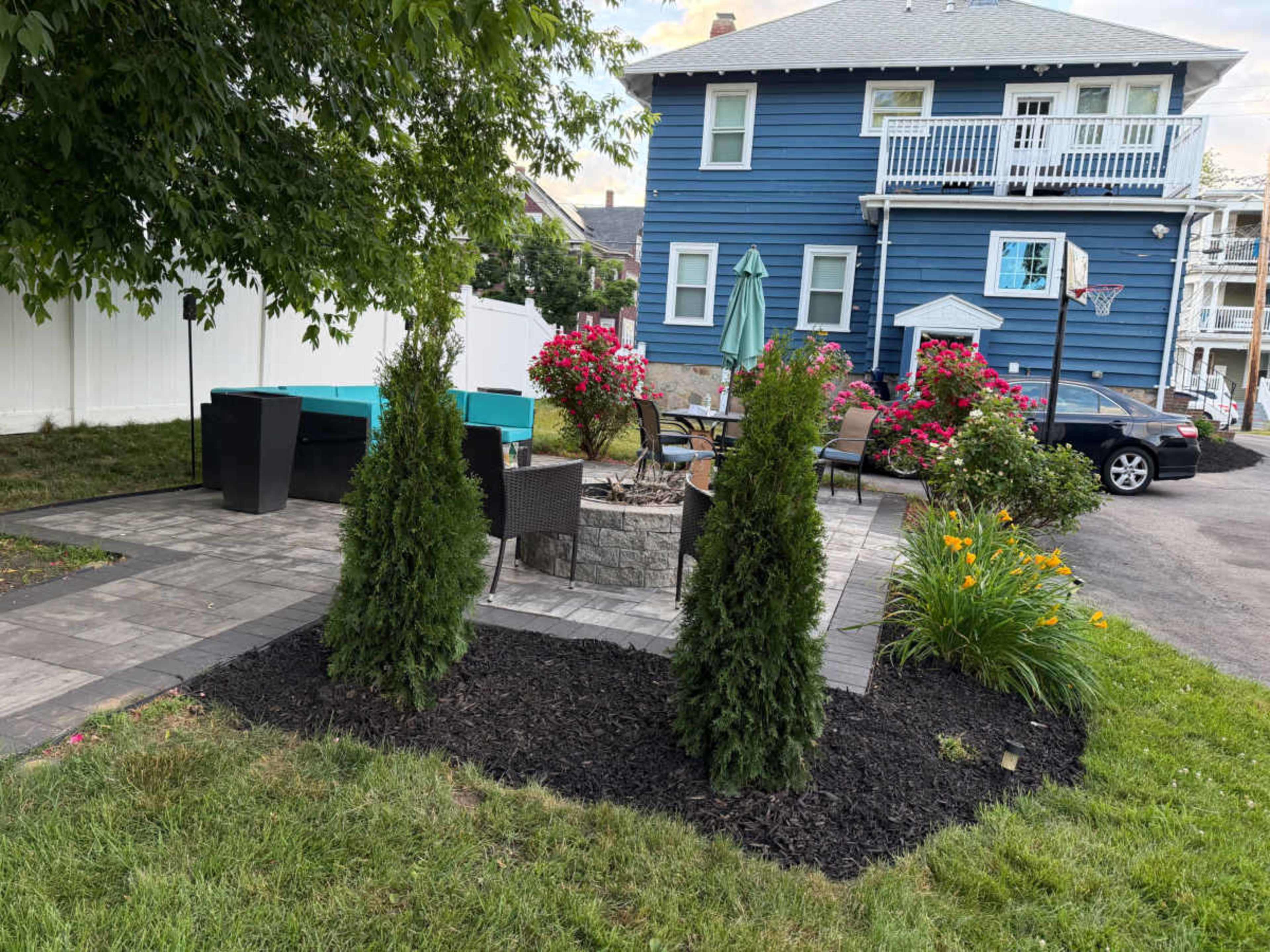 The image shows a landscaped outdoor patio area with seating surrounded by flower beds, two tall shrubs, and a blue house in the background.