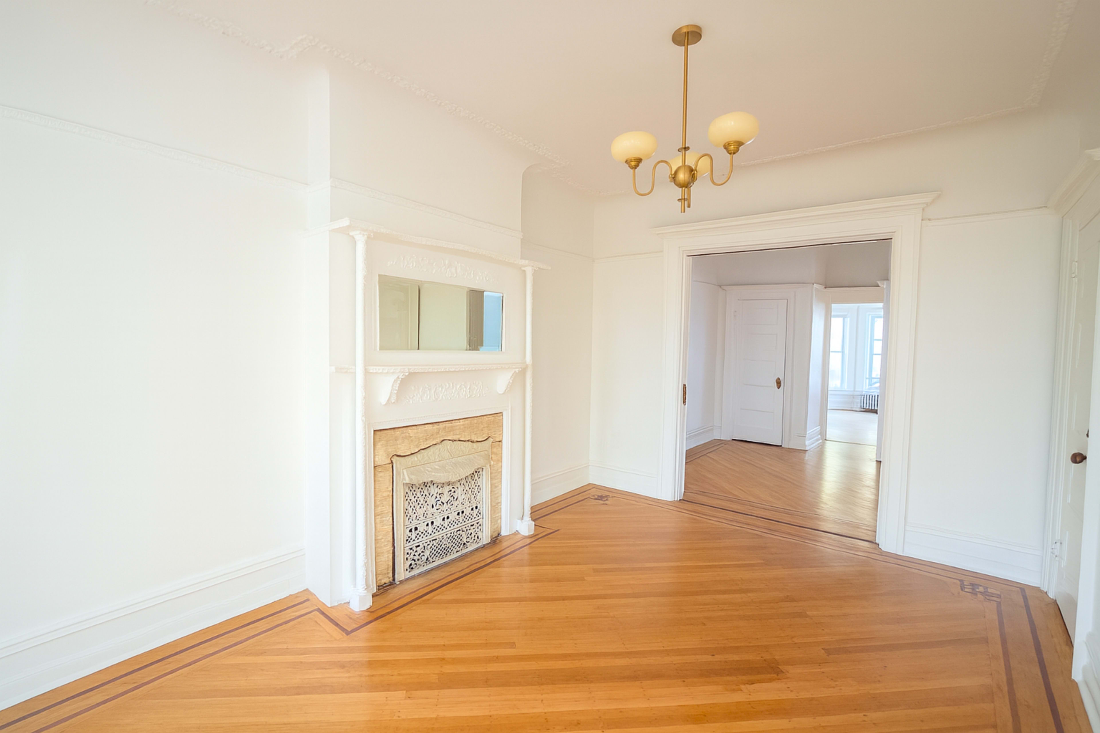 The image shows a bright, empty room with hardwood flooring, a decorative fireplace, and an adjacent doorway leading to another room.