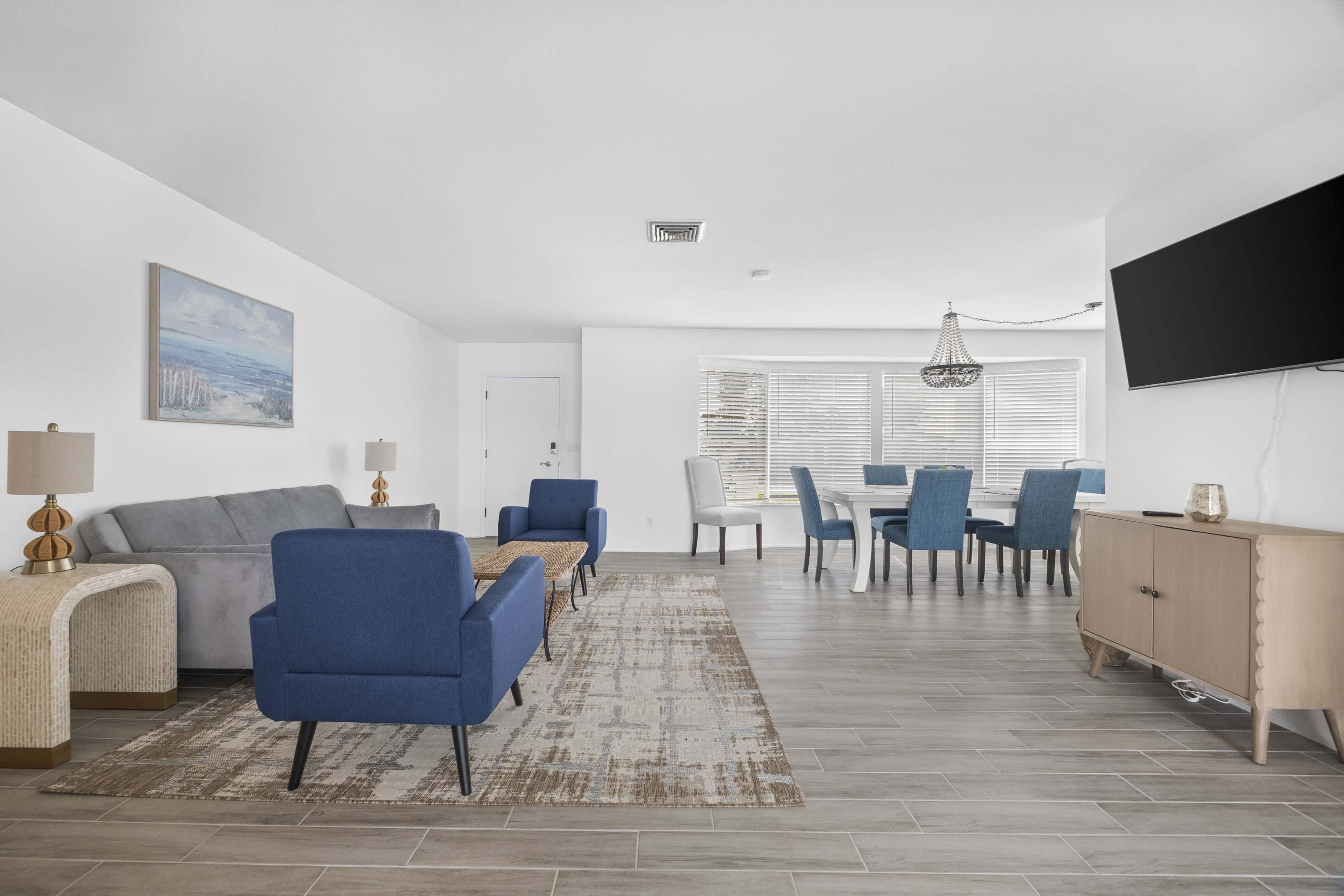 A modern living space with a gray sofa, two blue chairs, a dining area with a round table, and a television mounted on the wall.