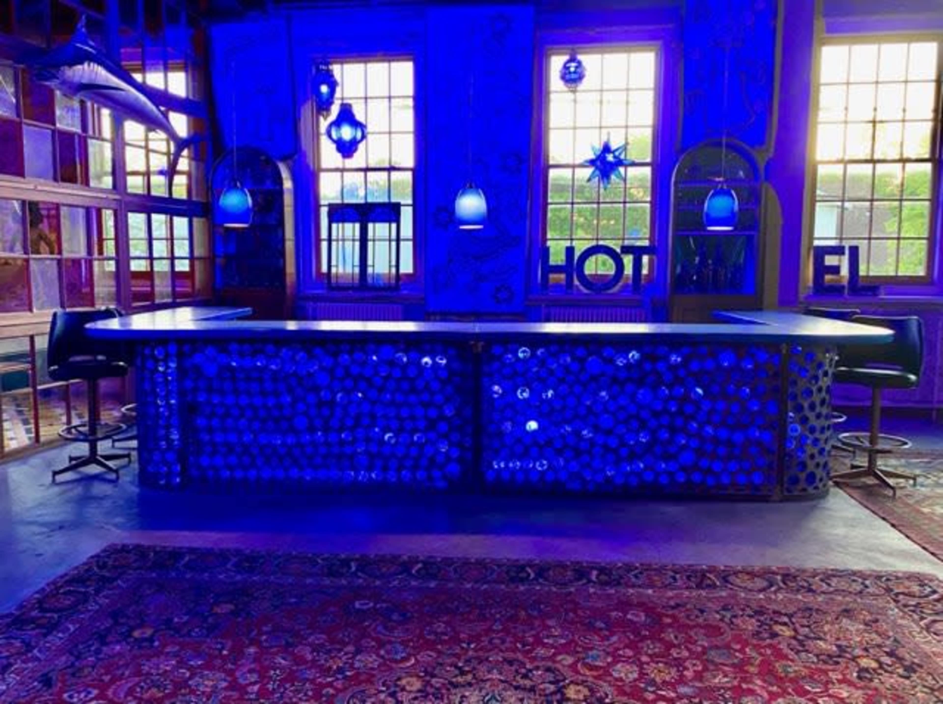A bar with a blue-lit counter and high stools is set against colorful walls and large windows.