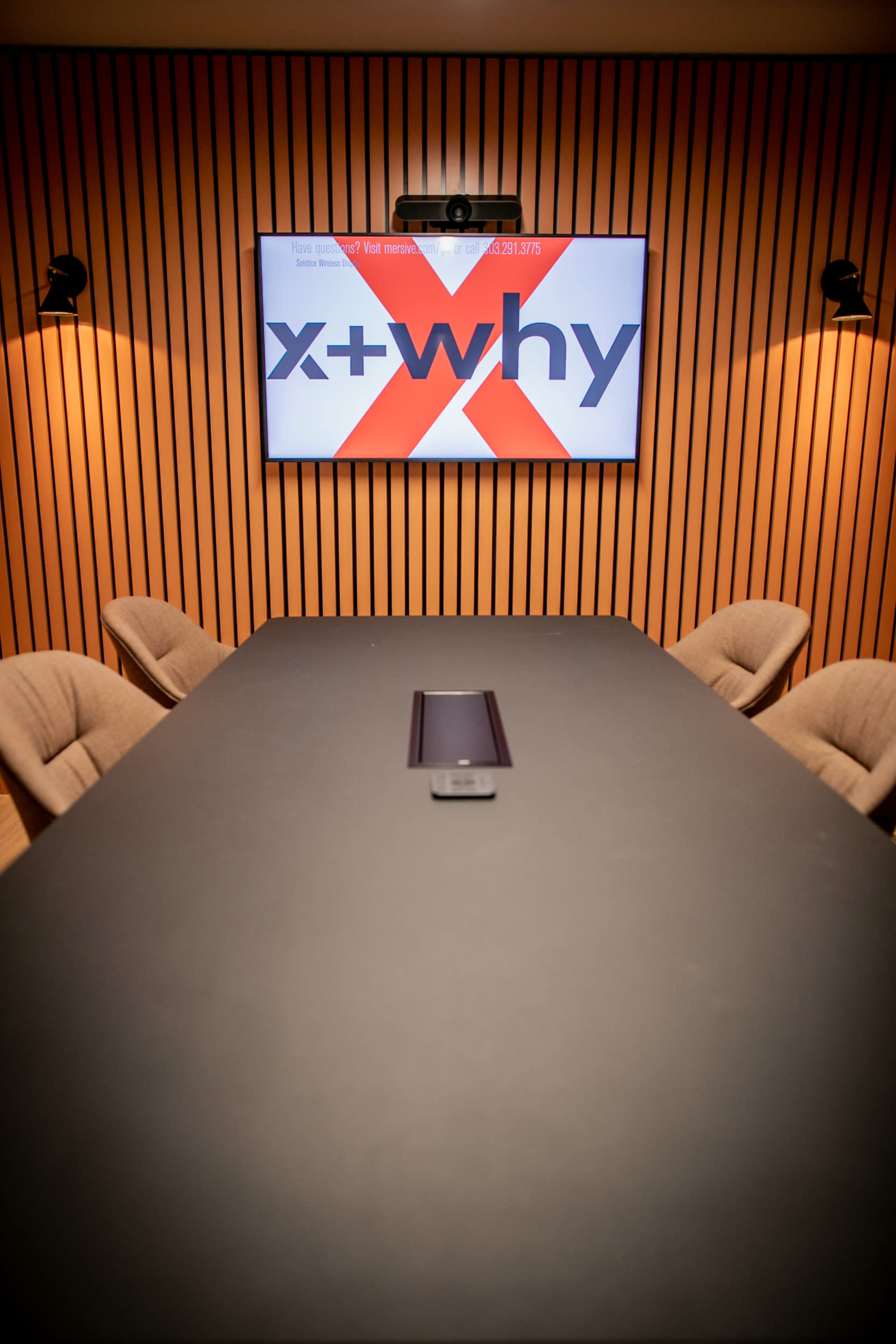A conference room features a long table with eight upholstered chairs and a large screen displaying the text "x + why" on a wooden-paneled wall.
