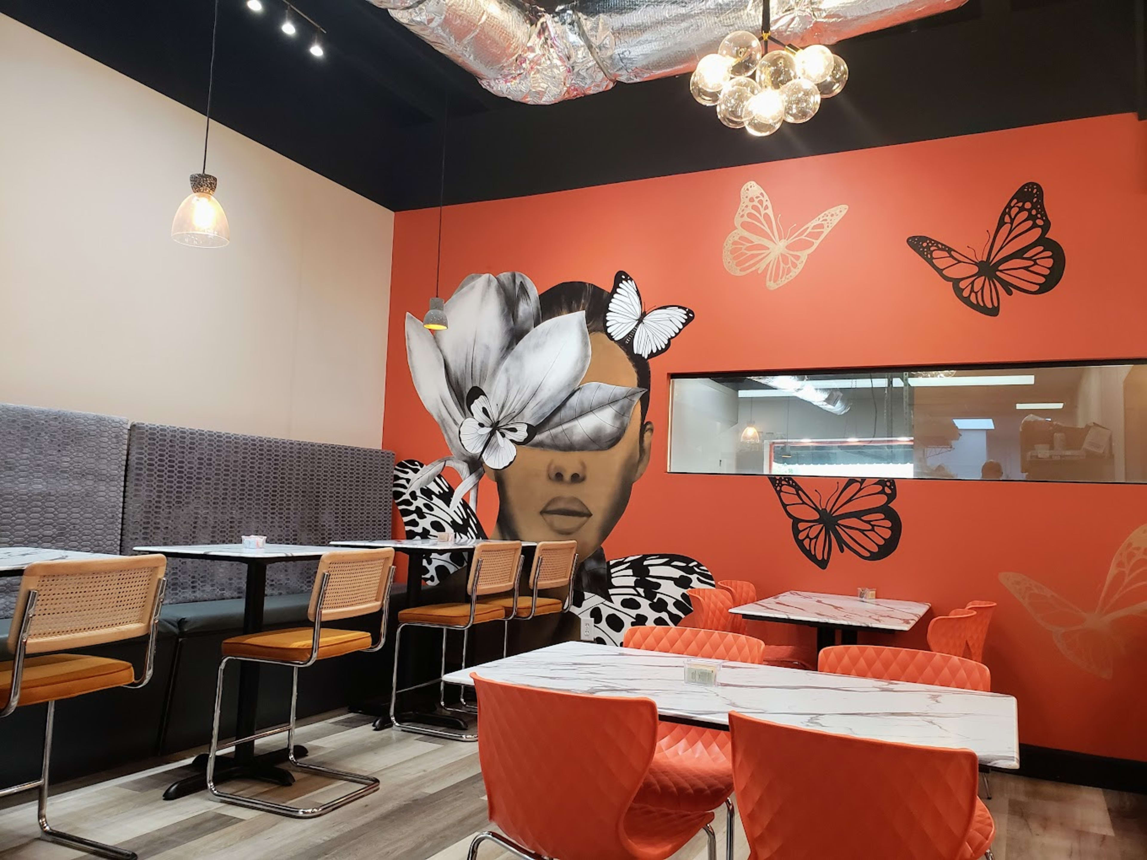 The image shows a modern restaurant interior featuring a large mural of a woman adorned with flowers and butterflies on an orange wall, alongside rows of tables and chairs.