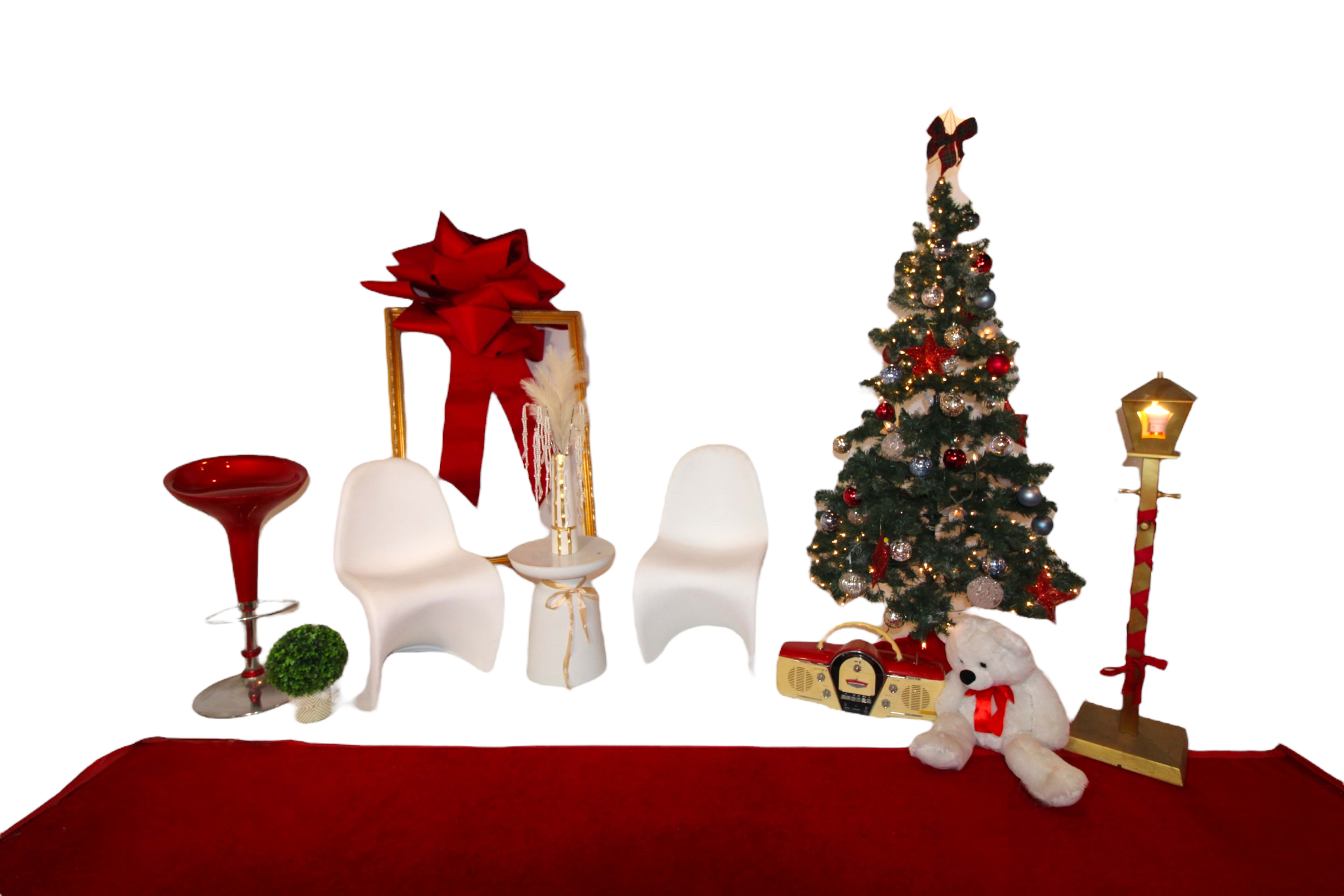 A festive arrangement with a decorated Christmas tree, two white chairs, a red backdrop, and various holiday-themed decorations, including a large bow and a teddy bear.