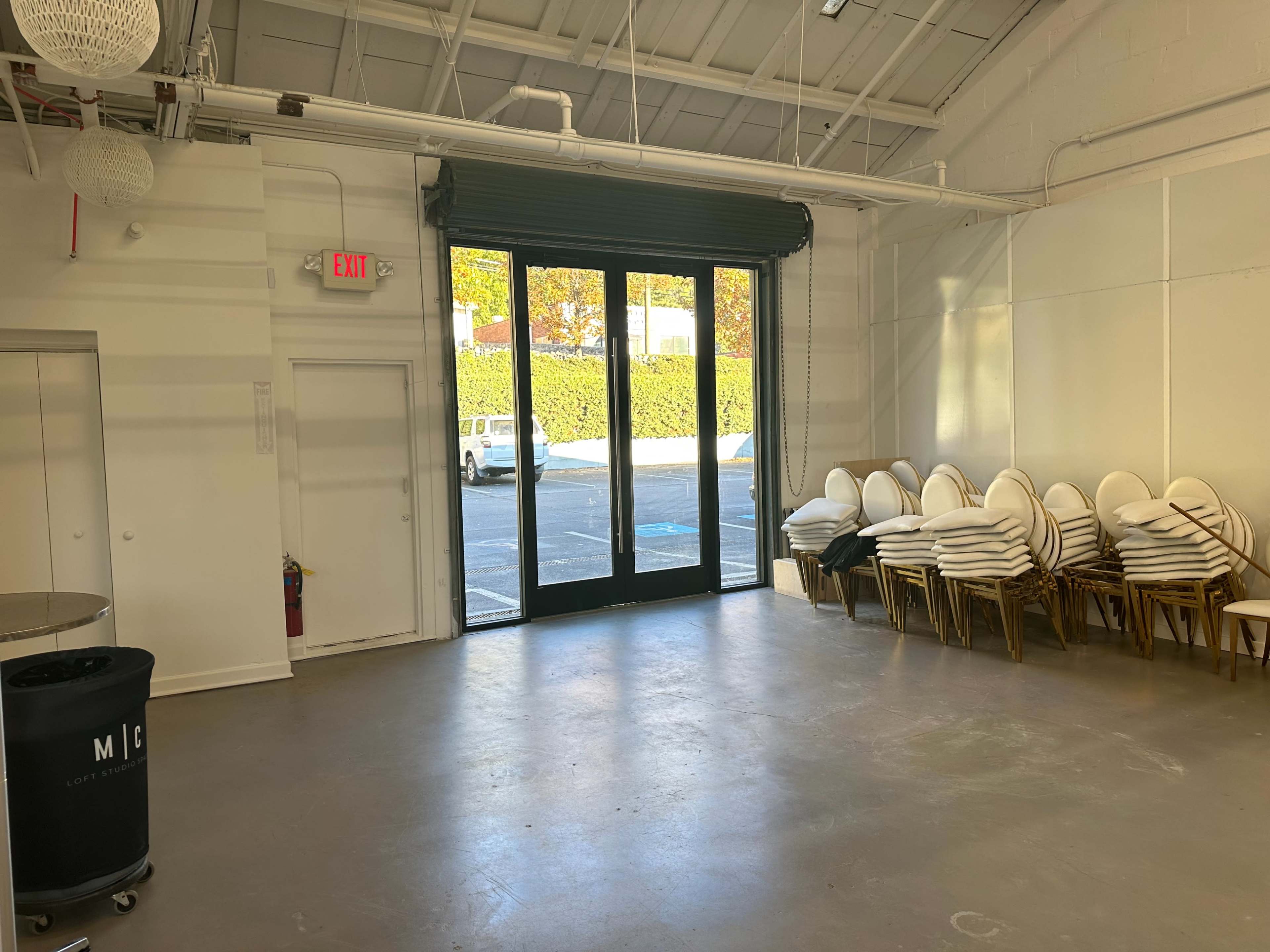 An empty room with a large set of double glass doors leading to an outdoor area, and stacks of white chairs against one wall.