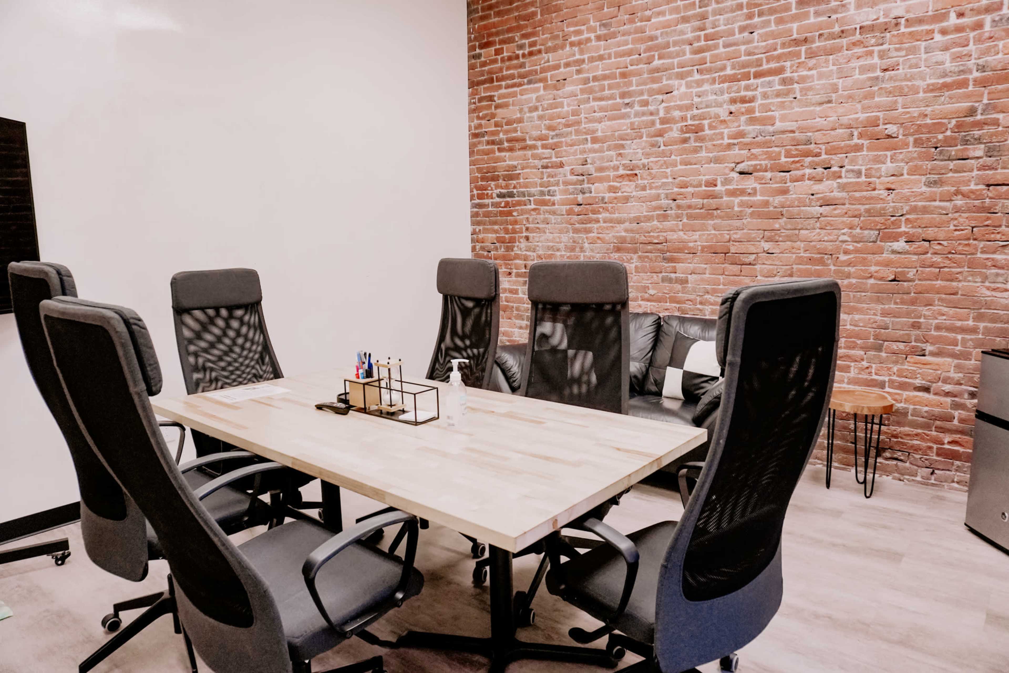 A conference room features a wooden table surrounded by six ergonomic chairs, with a brick wall backdrop and a leather couch in the corner.