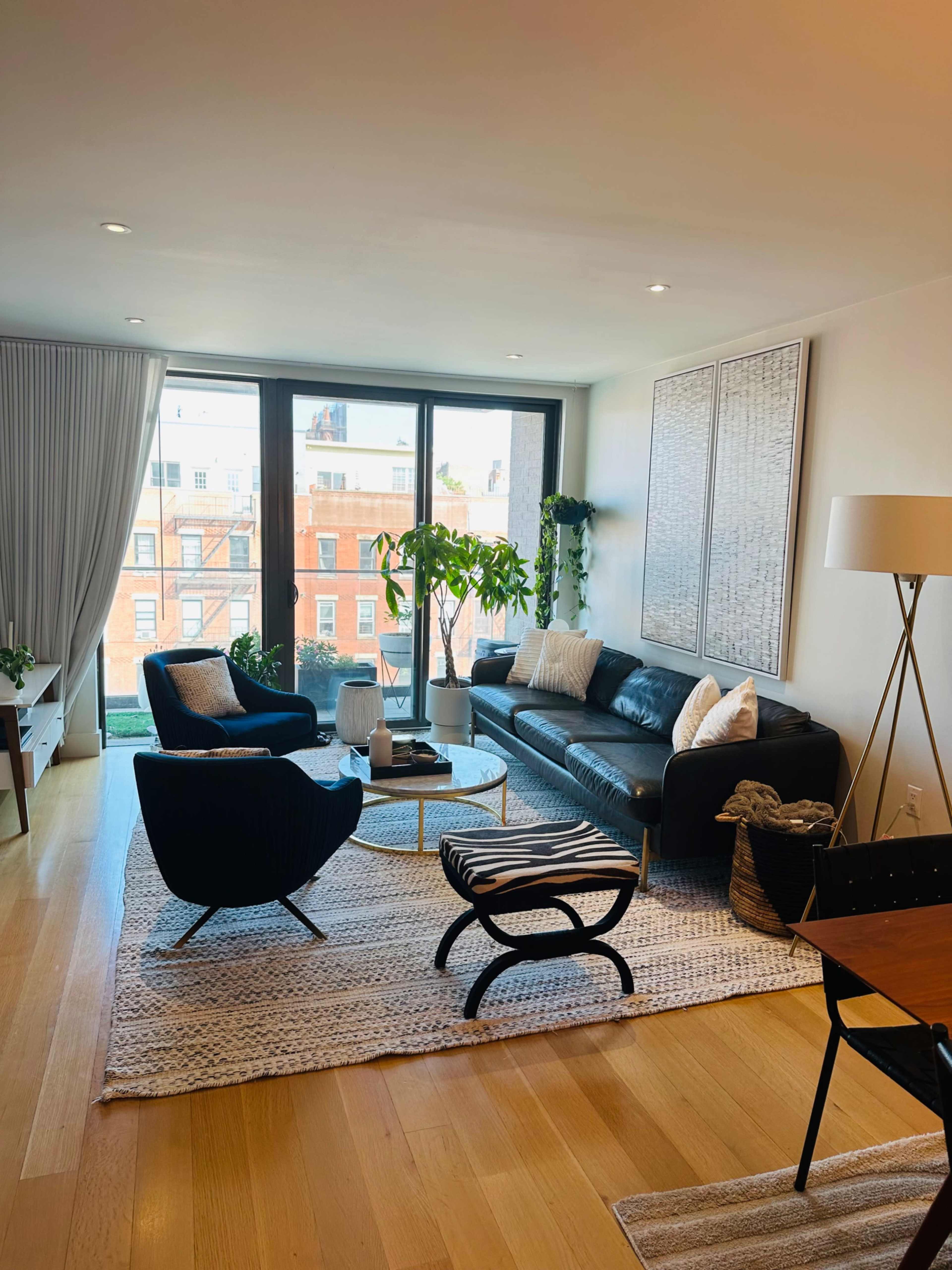 A modern living room with a black leather sofa, two armchairs, a coffee table, and large windows providing a view of city buildings outside.
