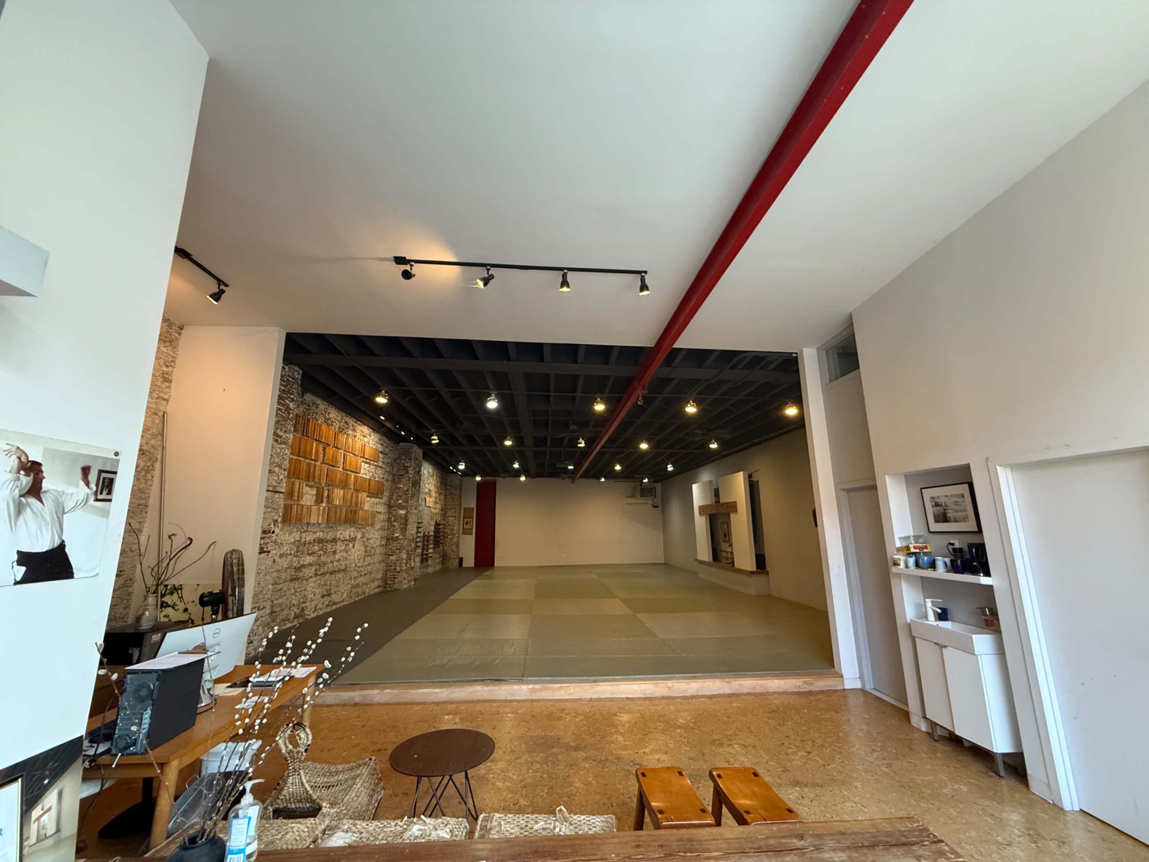 The image shows a spacious, empty studio with a large, open area featuring mats, wood paneling on the walls, and minimal furniture in the foreground.