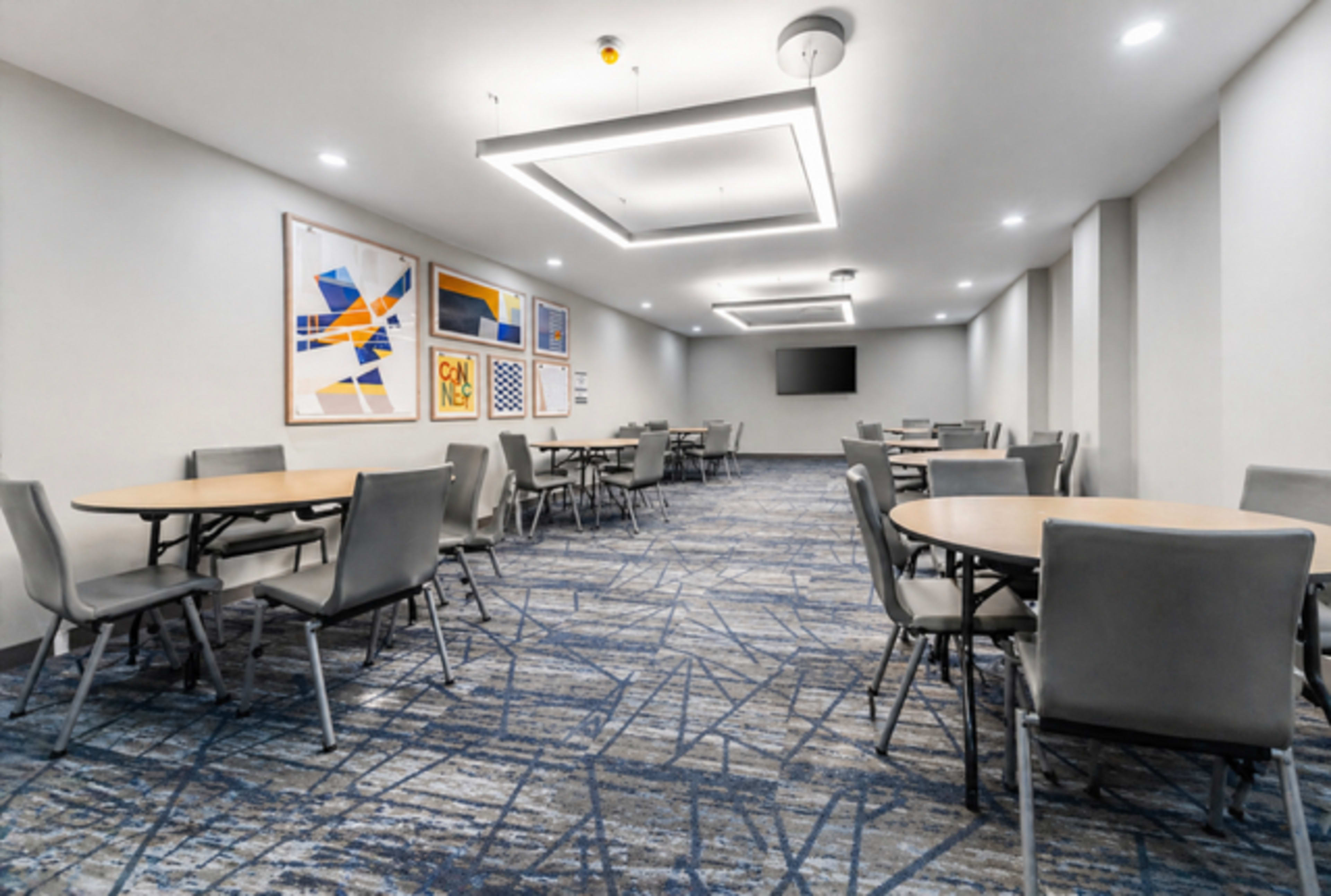 The image shows a modern meeting room with several circular tables, gray chairs, and abstract artwork on the walls.