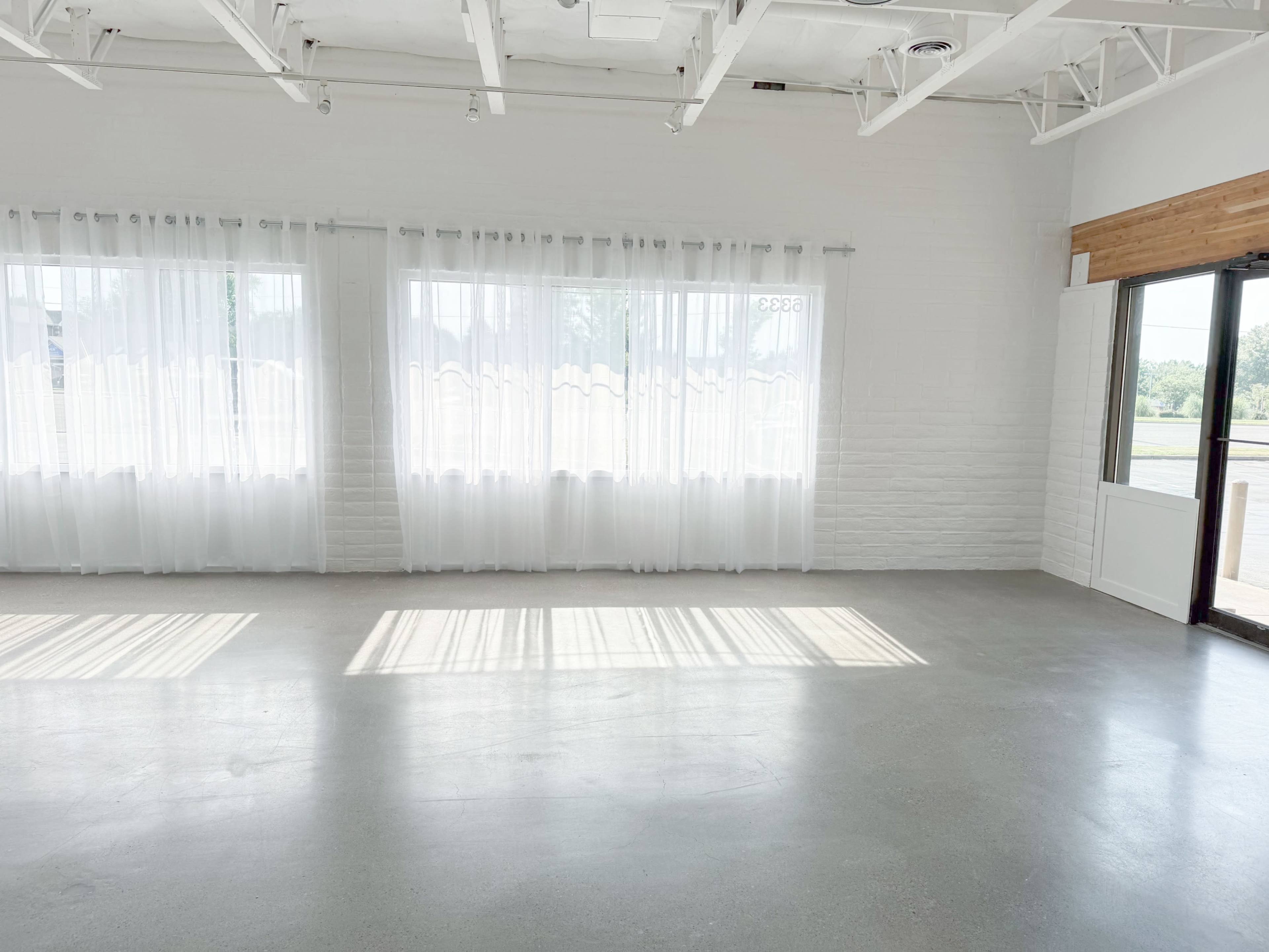 A spacious, bright room with large windows dressed in sheer white curtains and a polished concrete floor.