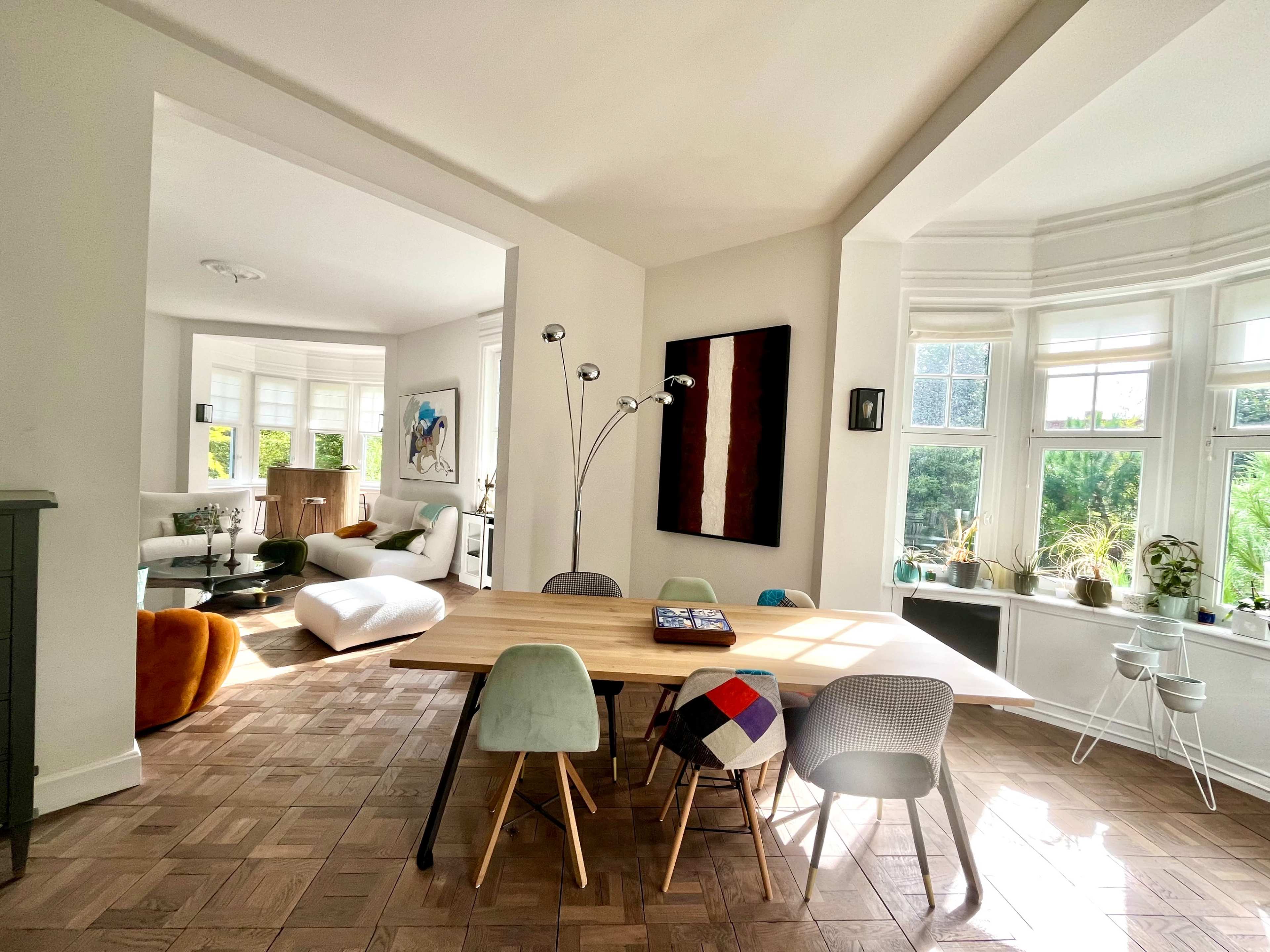 A spacious room features a wooden dining table surrounded by various chairs, with a living area visible in the background that includes a sofa and artwork.