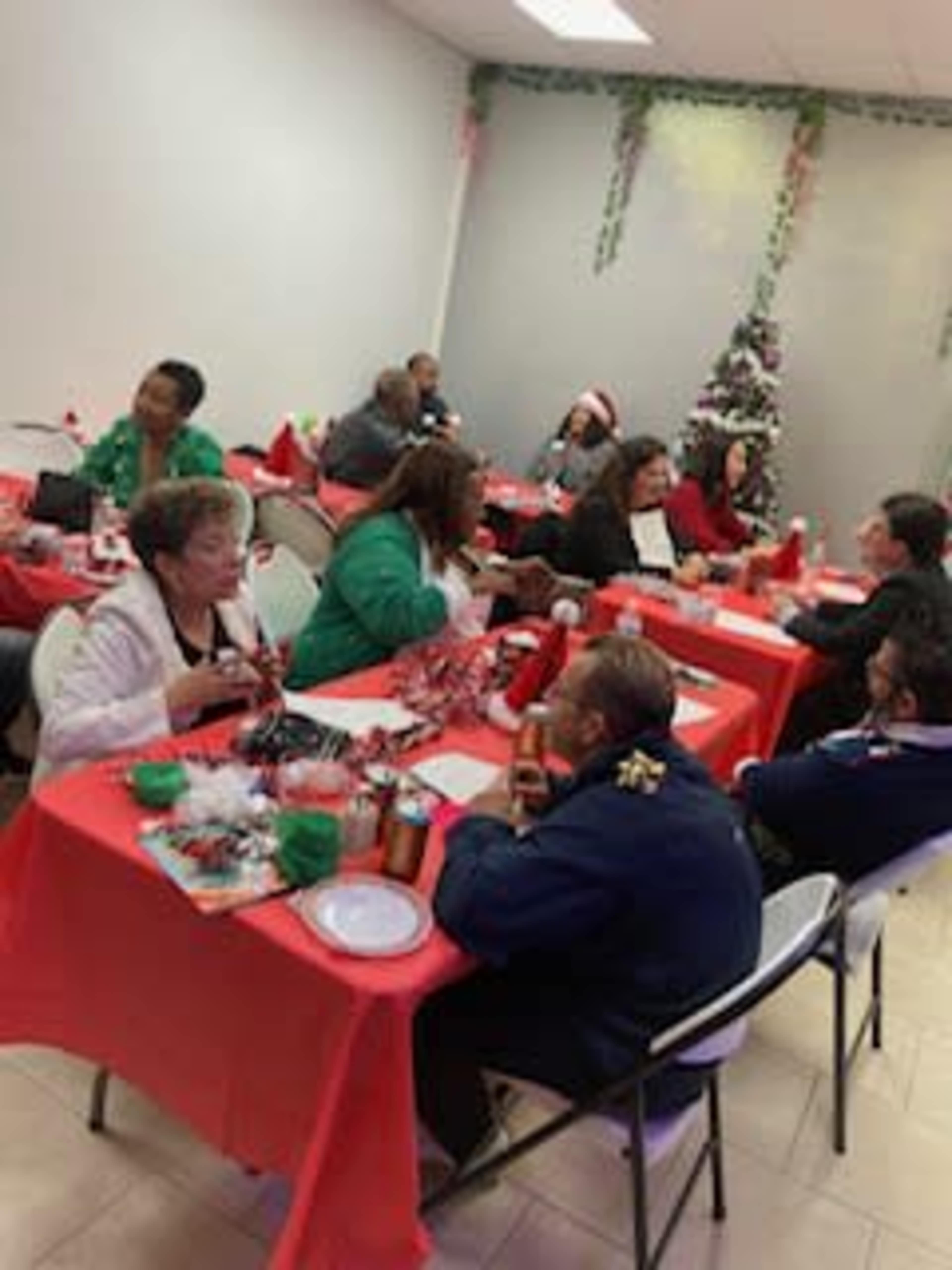 A group of people sits around tables adorned with red tablecloths, enjoying a festive gathering in a decorated room with a Christmas tree.