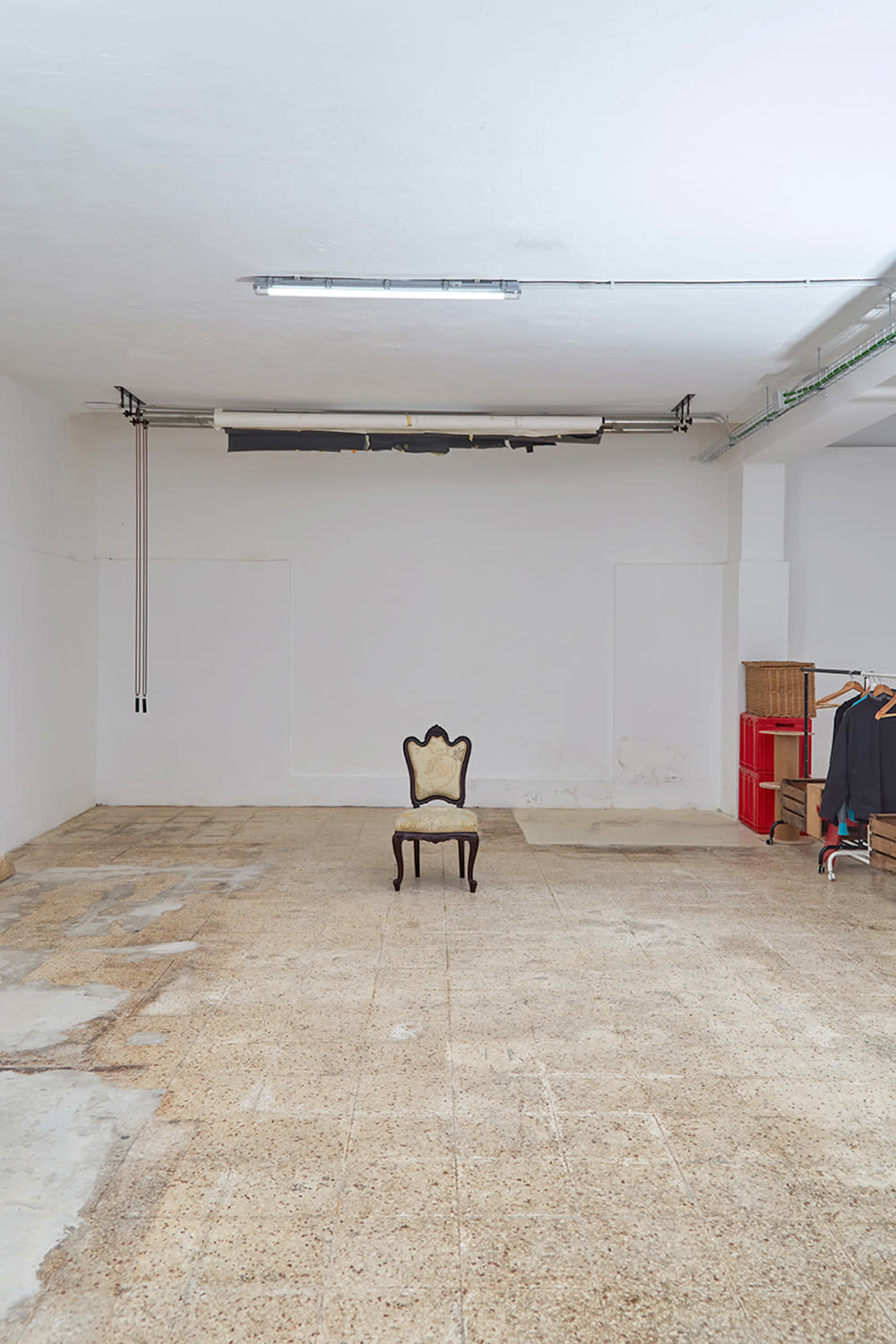 An empty room features a single ornate chair on a bare floor, with clothing hanging on a rack in the background.