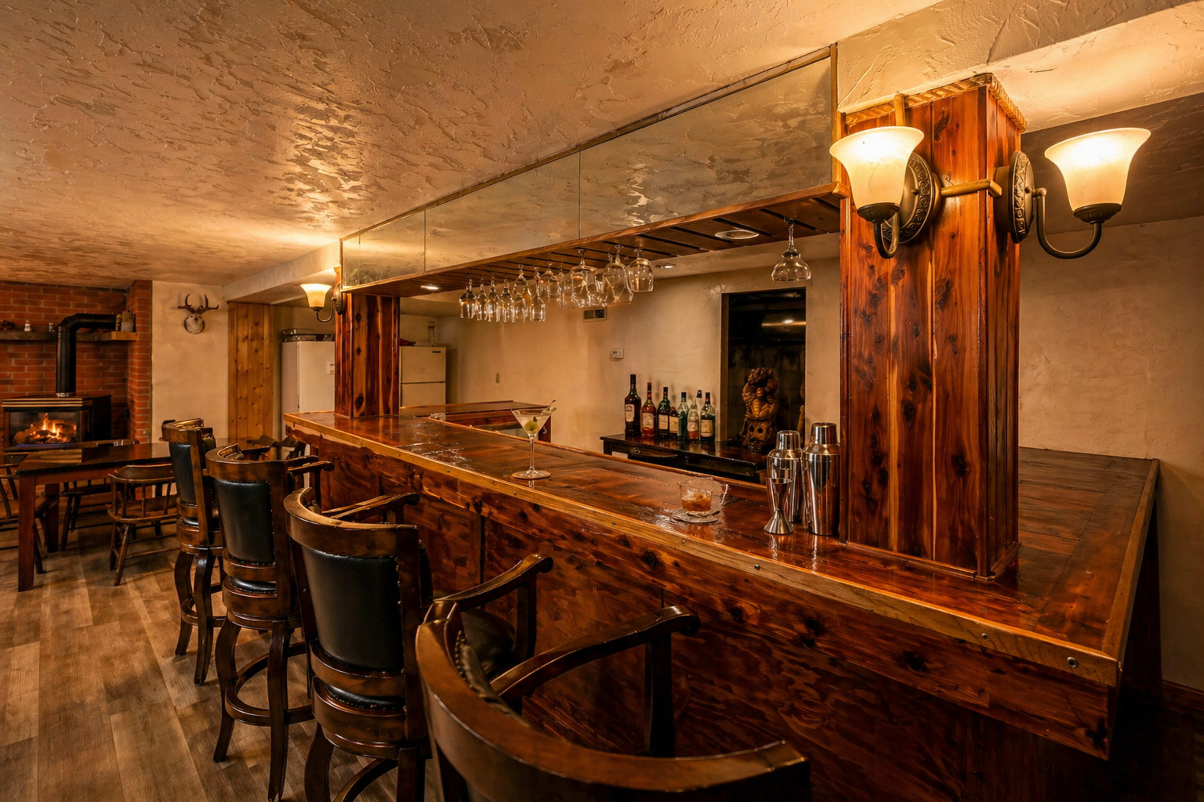 The image shows a wooden bar area with several high stools, a mirror reflecting the bar's backlit shelves of liquor, and a cozy fireplace in the background.