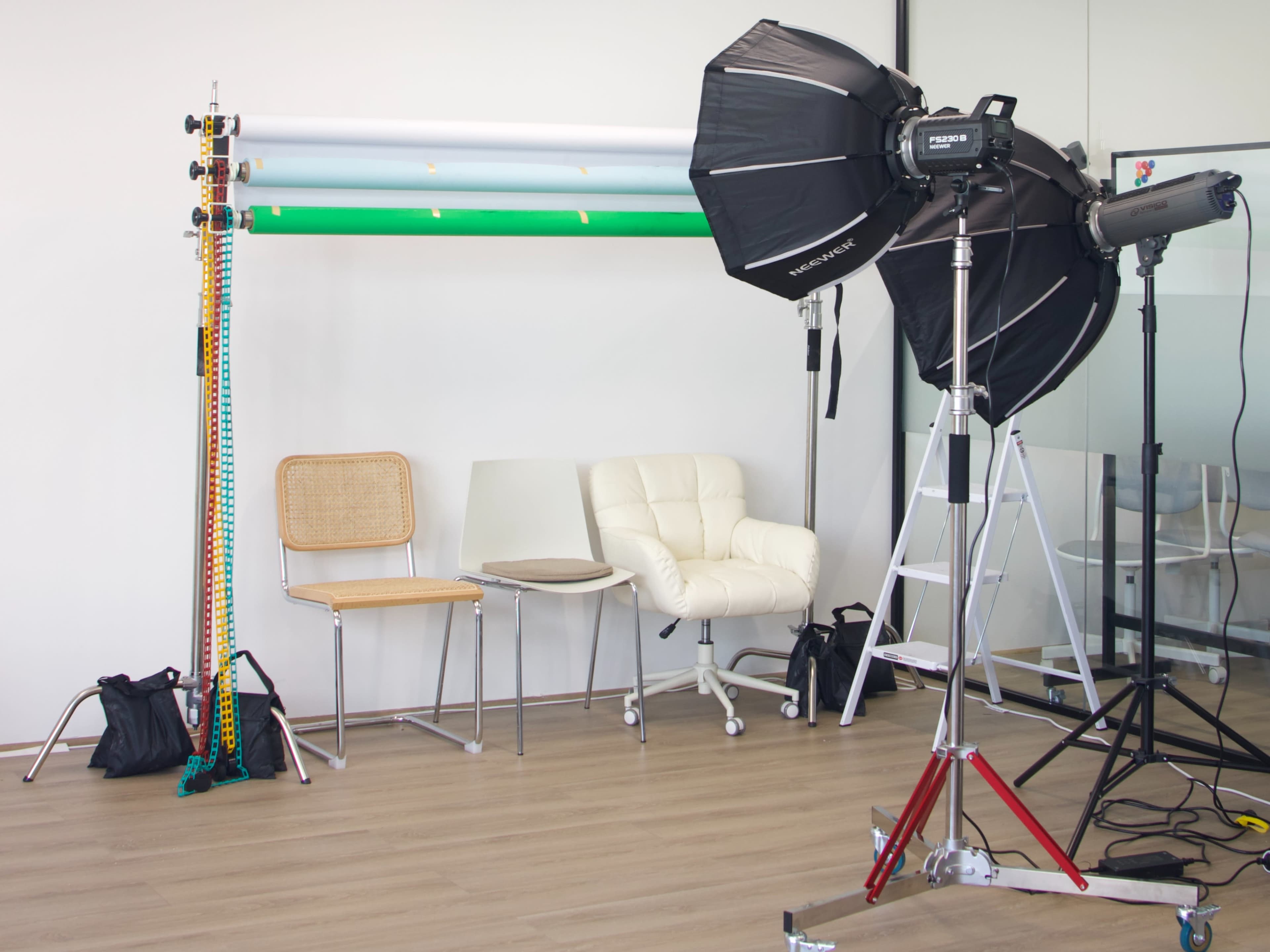 The image shows a photography setup in a studio featuring a light stand, backdrop rolls, various chairs, and equipment on a wooden floor.