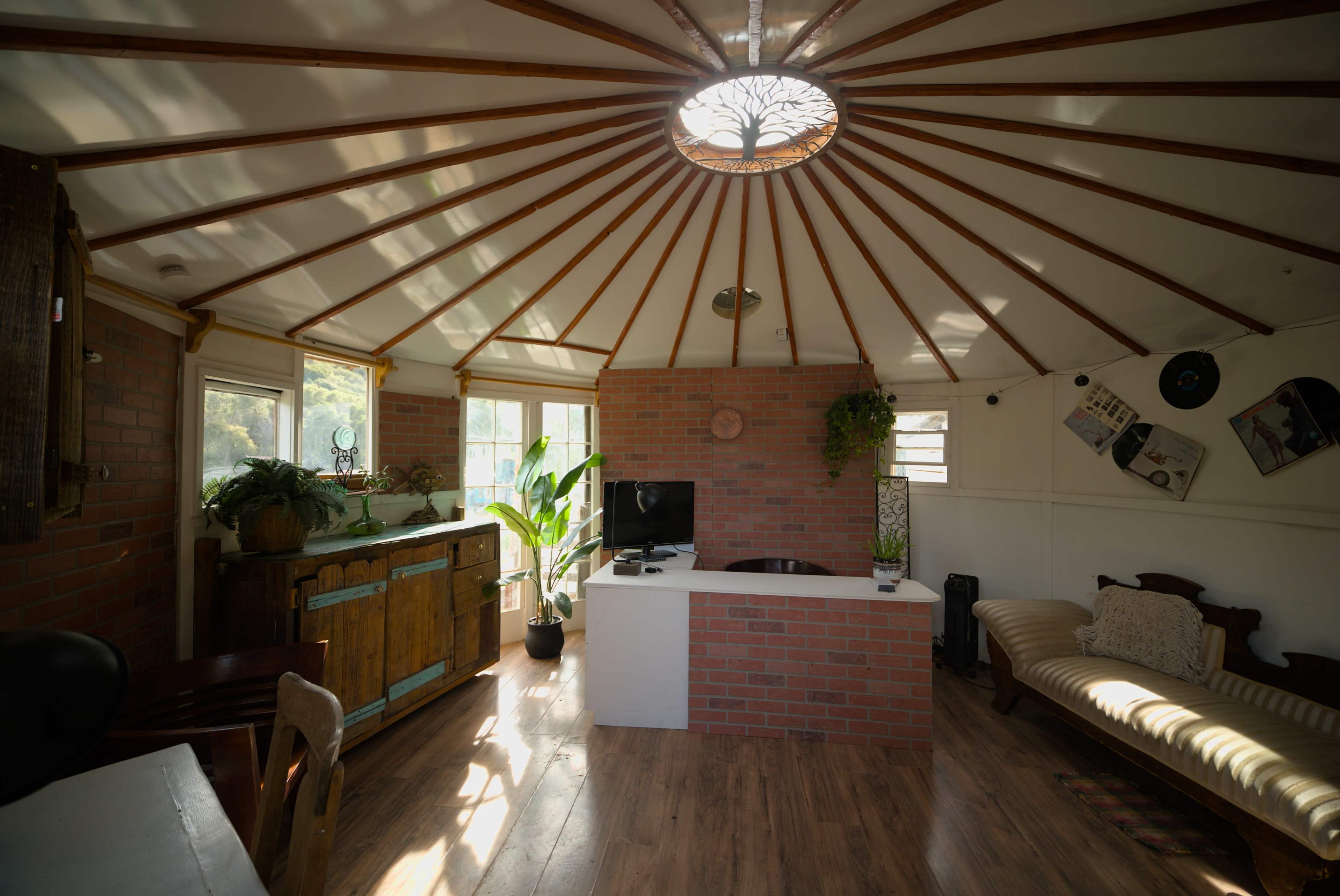 The interior of a circular yurt features wooden beams radiating from a central skylight, with a brick accent wall, a small kitchen area, and a seating space.