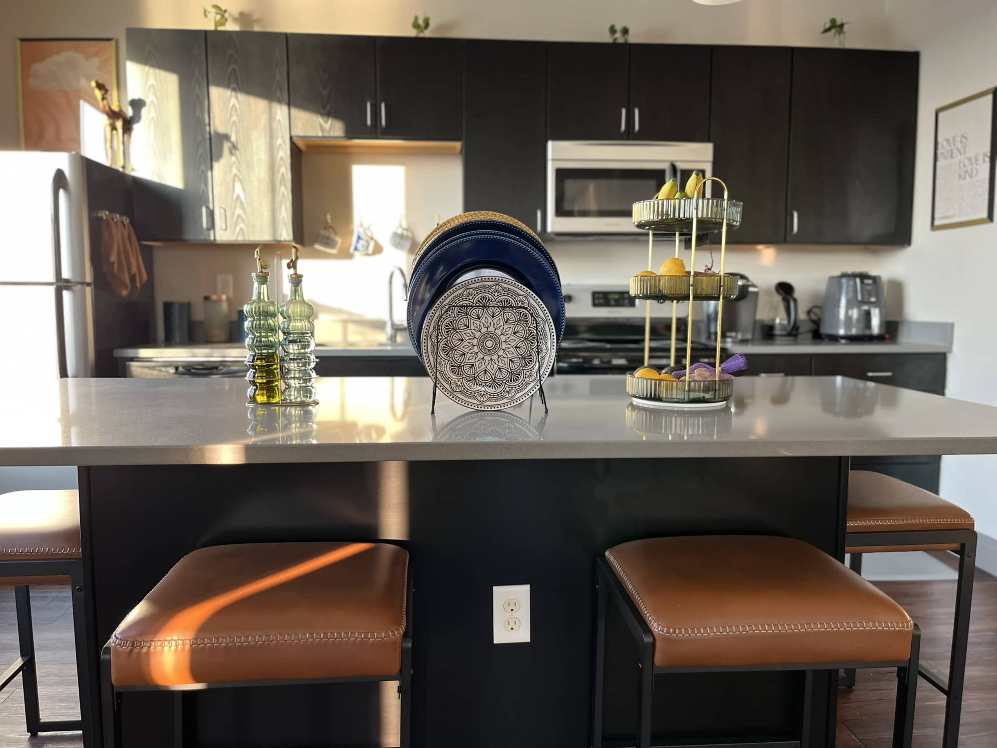 The image shows a modern kitchen with a central island, featuring a decorative plate, oils in glass bottles, and a tiered fruit stand, along with leather bar stools.