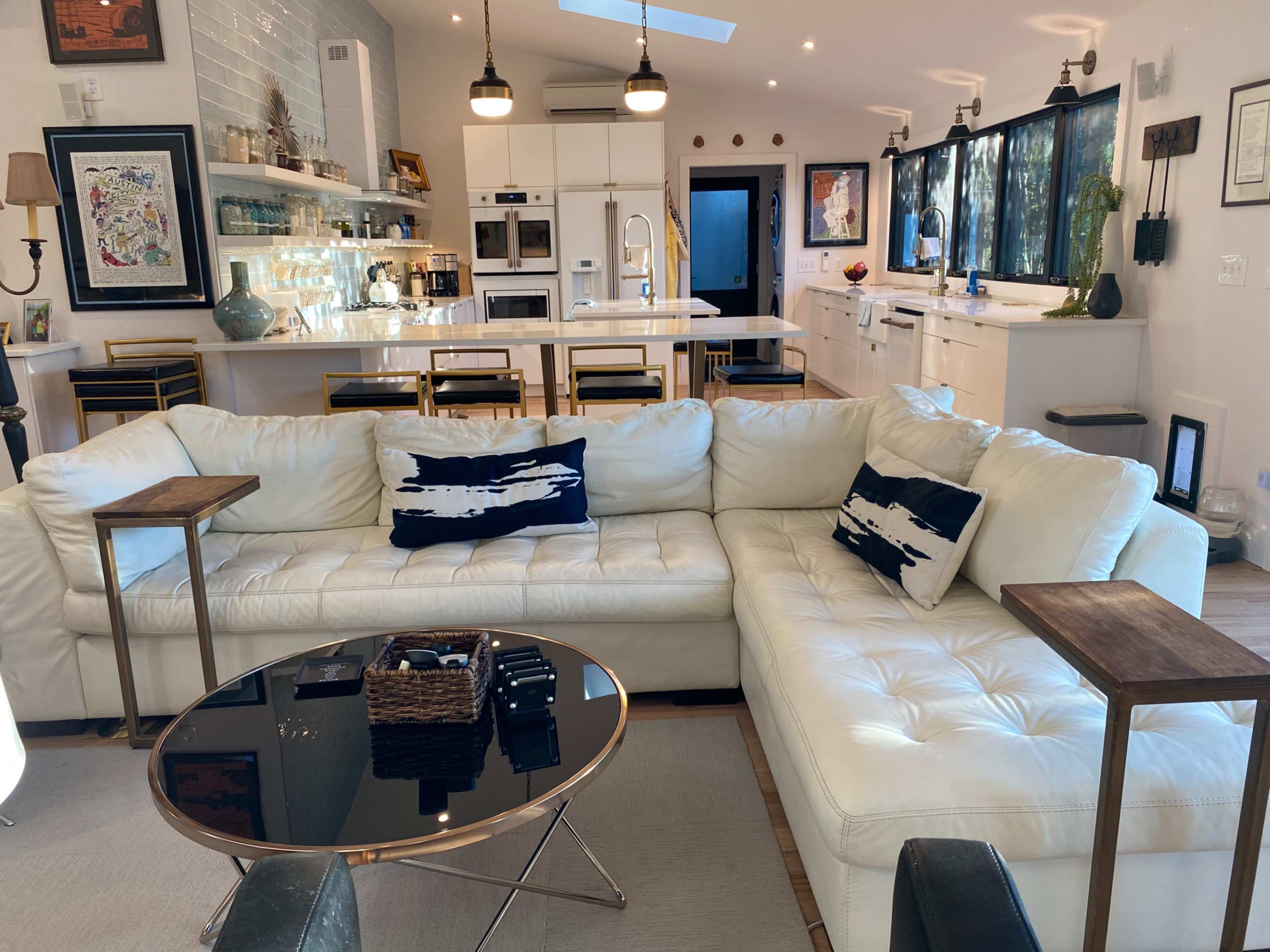 The image shows a modern, open-concept living area featuring a white sectional sofa, a black coffee table with a woven basket, and a kitchen in the background with white cabinetry and appliances.