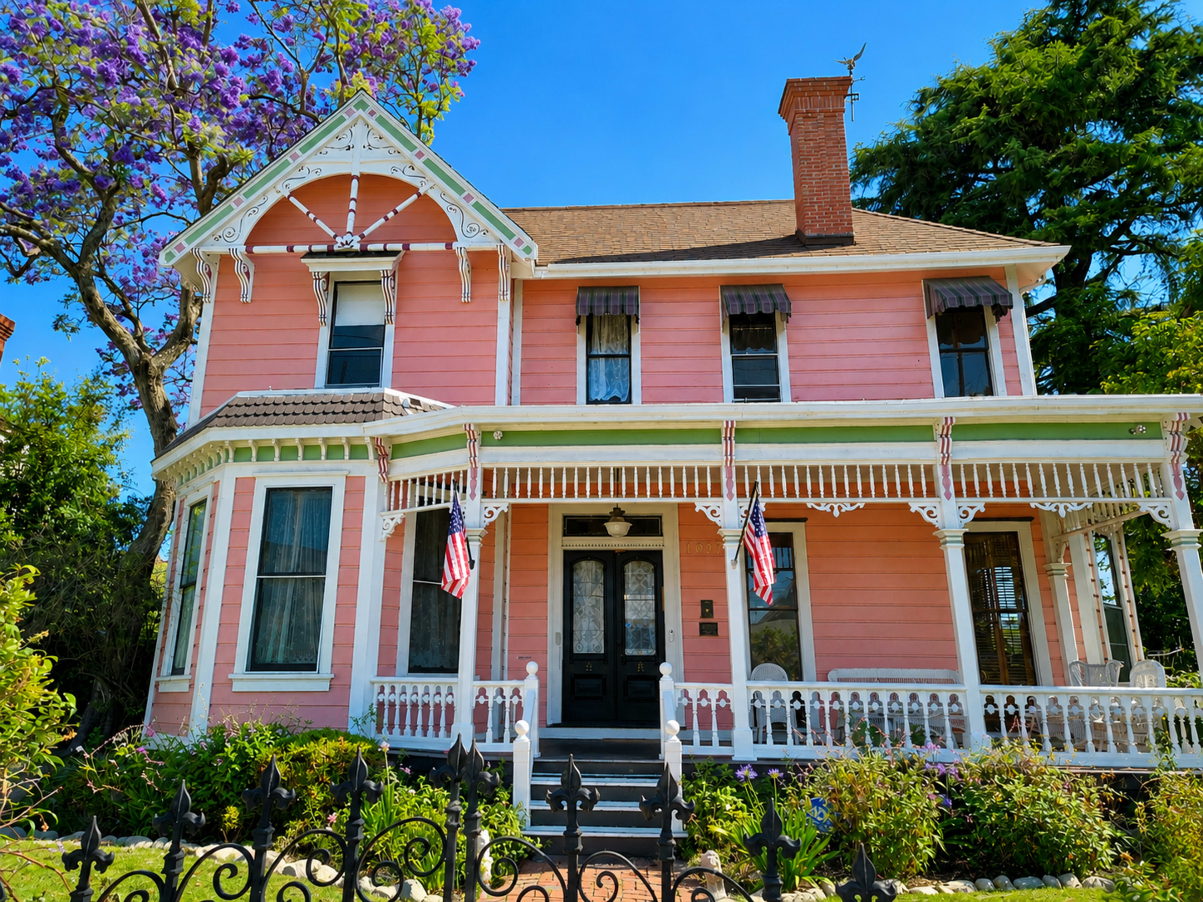 The image shows a two-story, pink Victorian-style house with white trim, featuring a front porch adorned with decorative railings and two American flags.