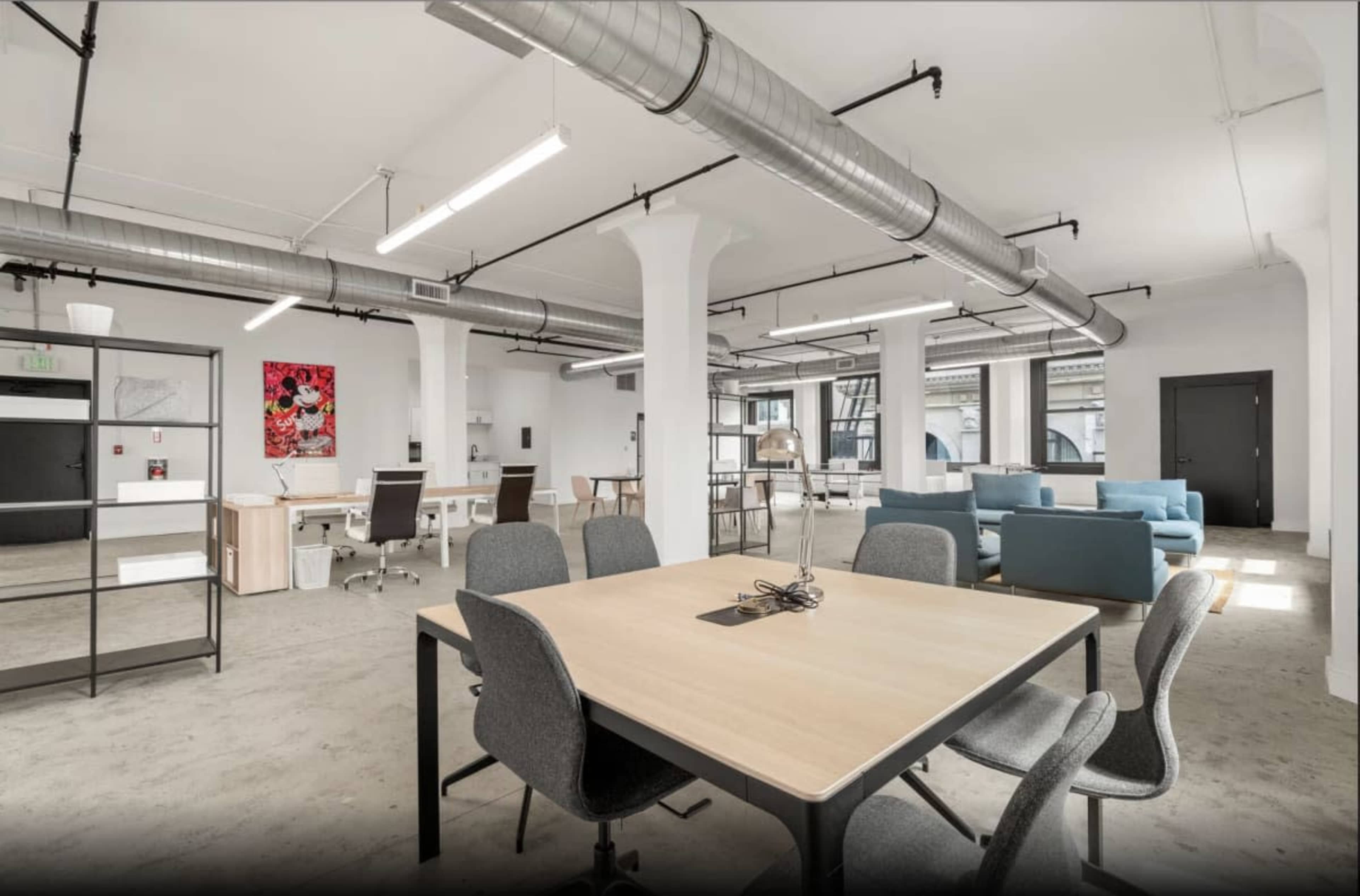 The image shows a spacious, modern office environment with a central wooden table surrounded by gray chairs, and various workstations and a lounge area in the background.