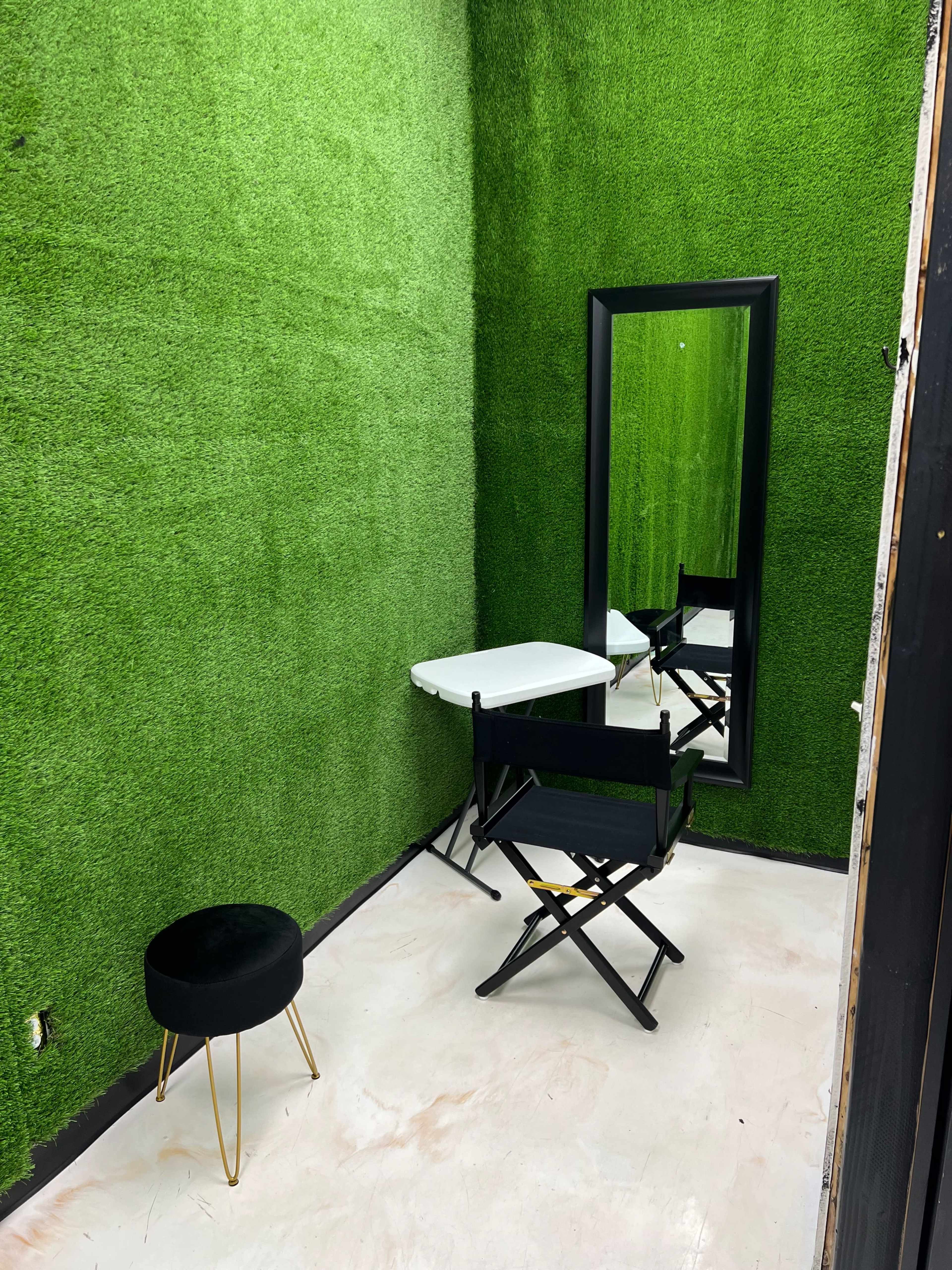 A small interior space with green grass-like walls, a mirror, a white table, a black stool, and a director's chair.