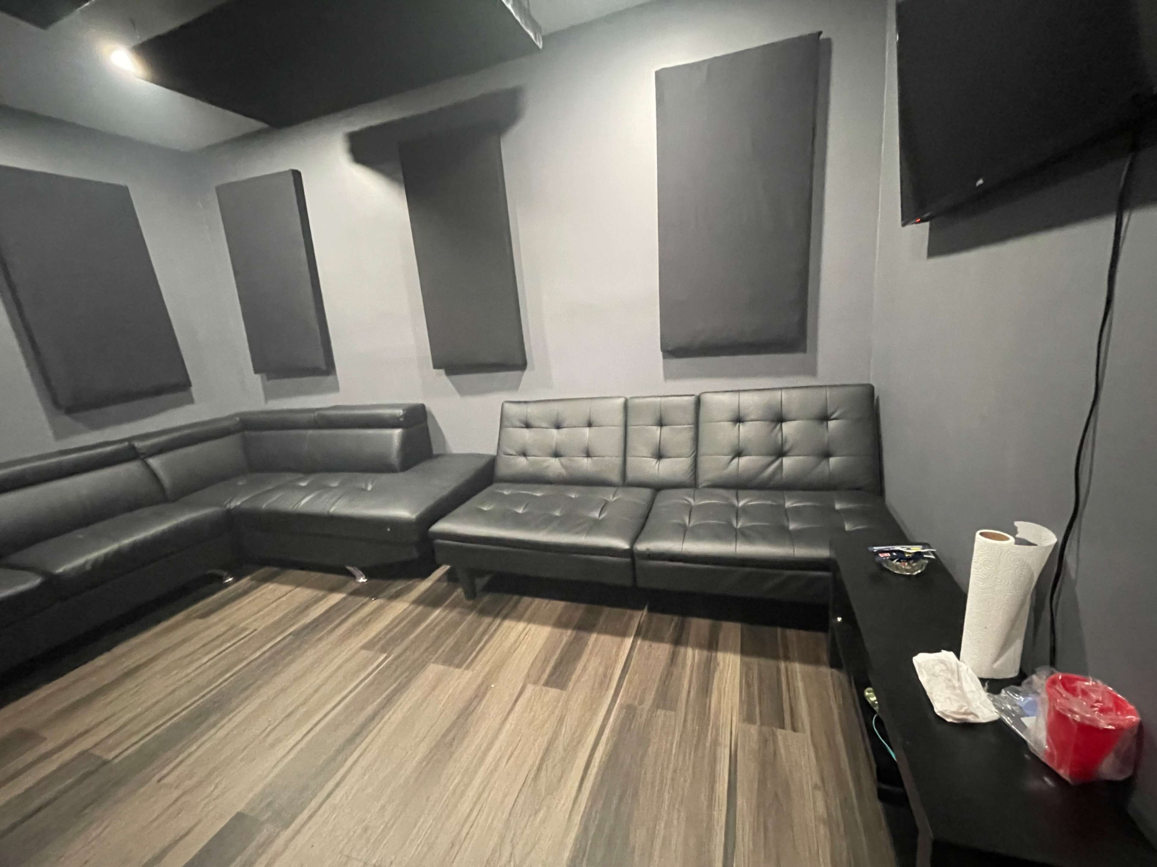 The image shows a room with dark gray walls, a black sectional couch, and acoustic panels on the walls, along with a television mounted in one corner.