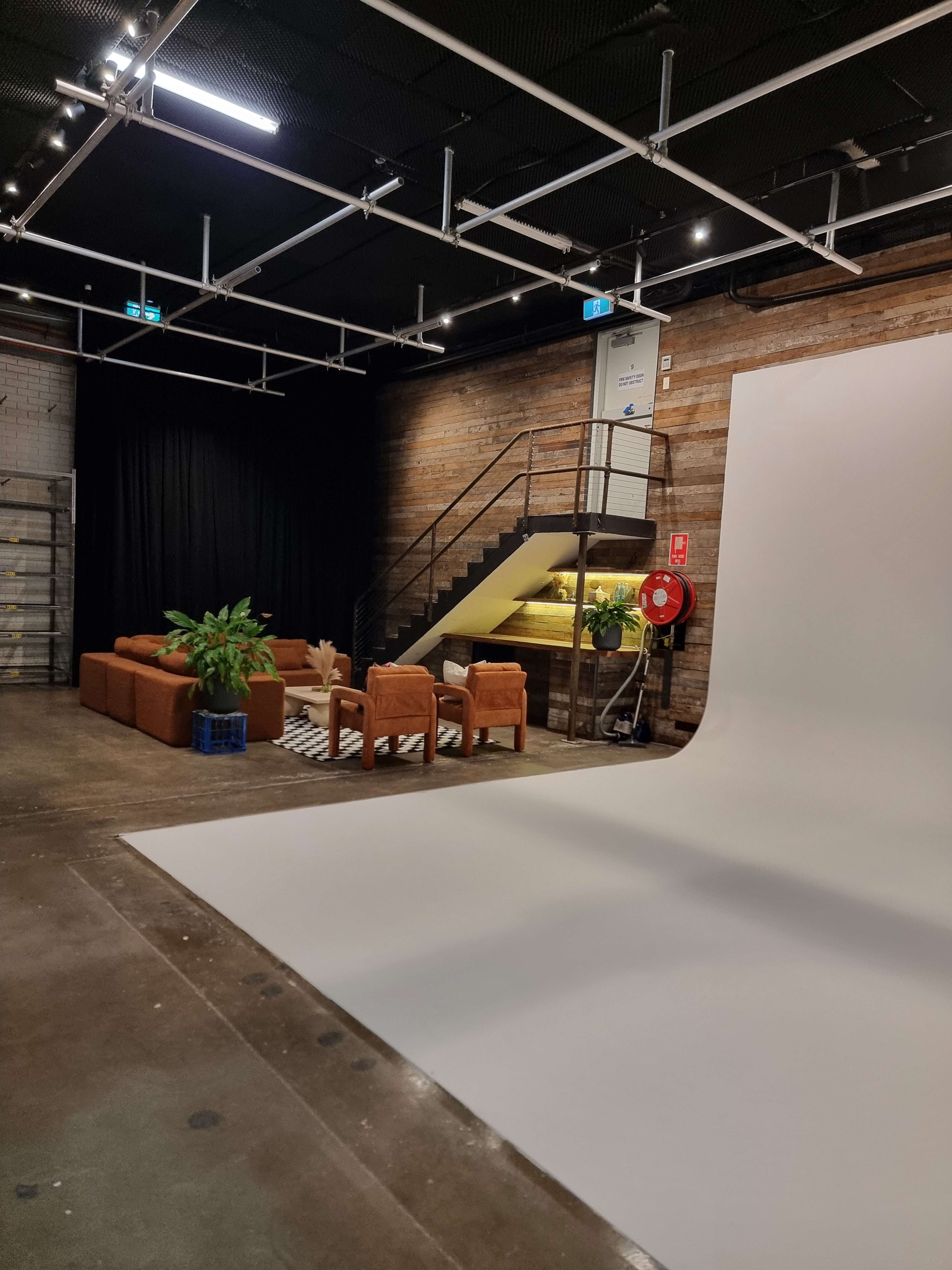 The image shows a spacious studio with a white backdrop, a brown sofa set, a staircase, and a plant, along with a black curtain in the background.