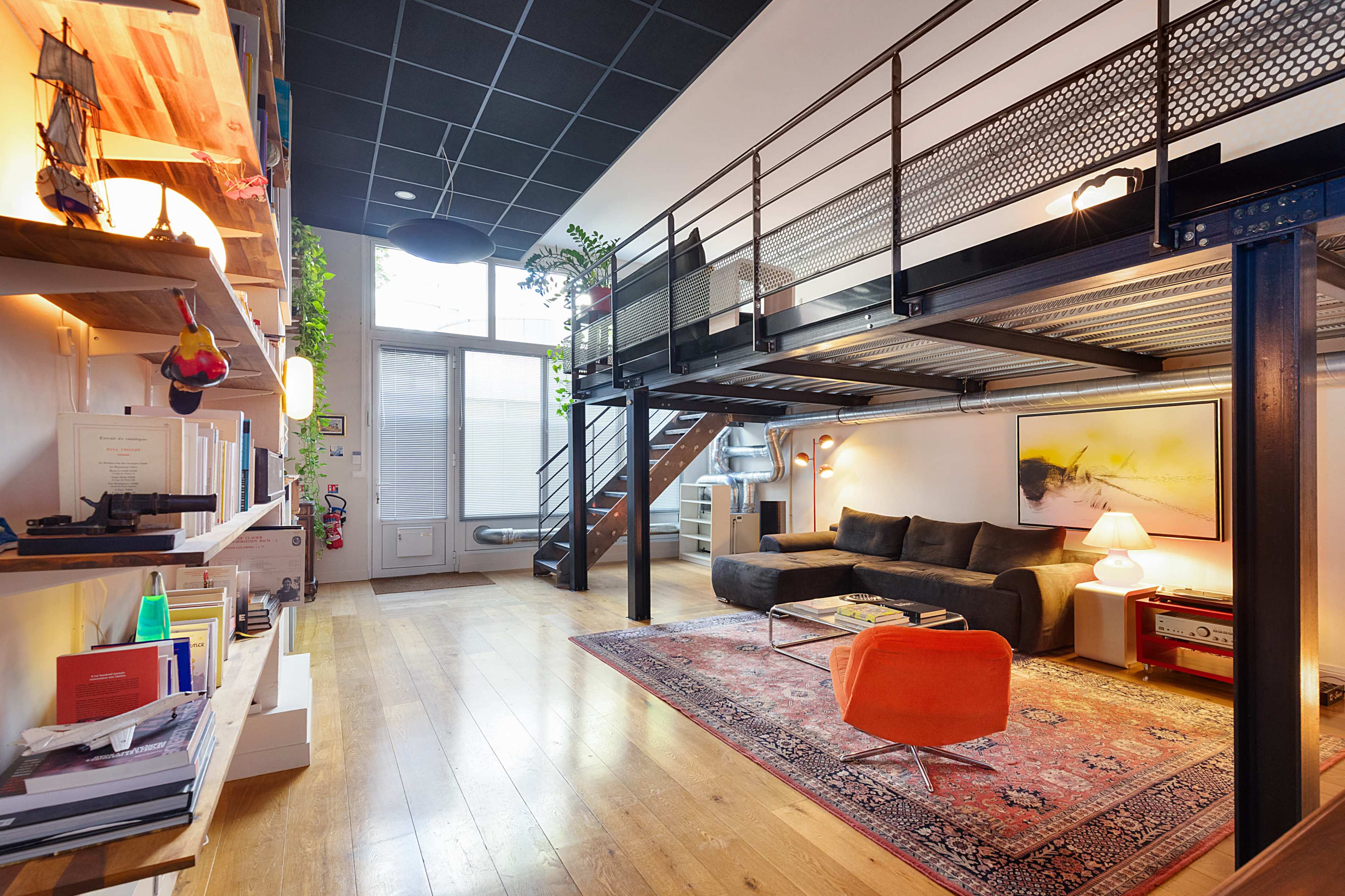 The image shows a modern, multi-level living space featuring a lofted area with a black metal railing, a vibrant orange chair, and a cozy seating arrangement on a wooden floor.