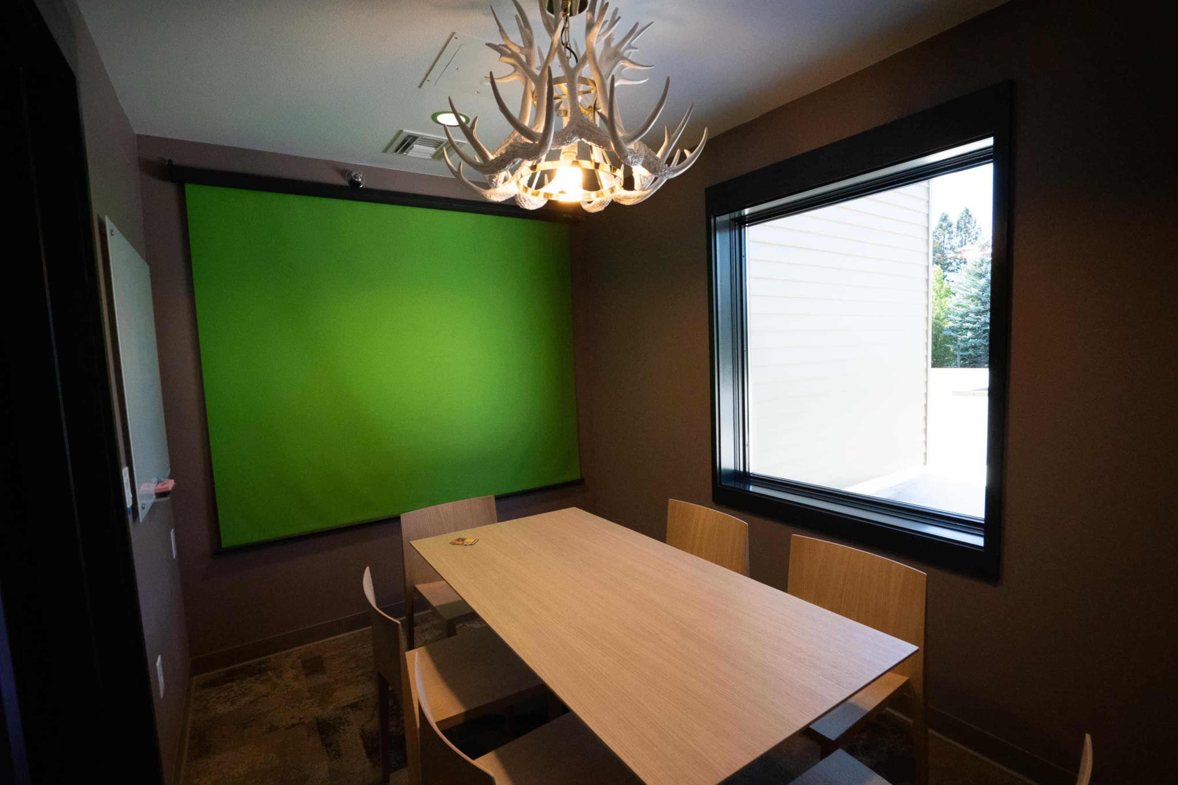 A meeting room features a wooden table surrounded by chairs and a green screen mounted on the wall.