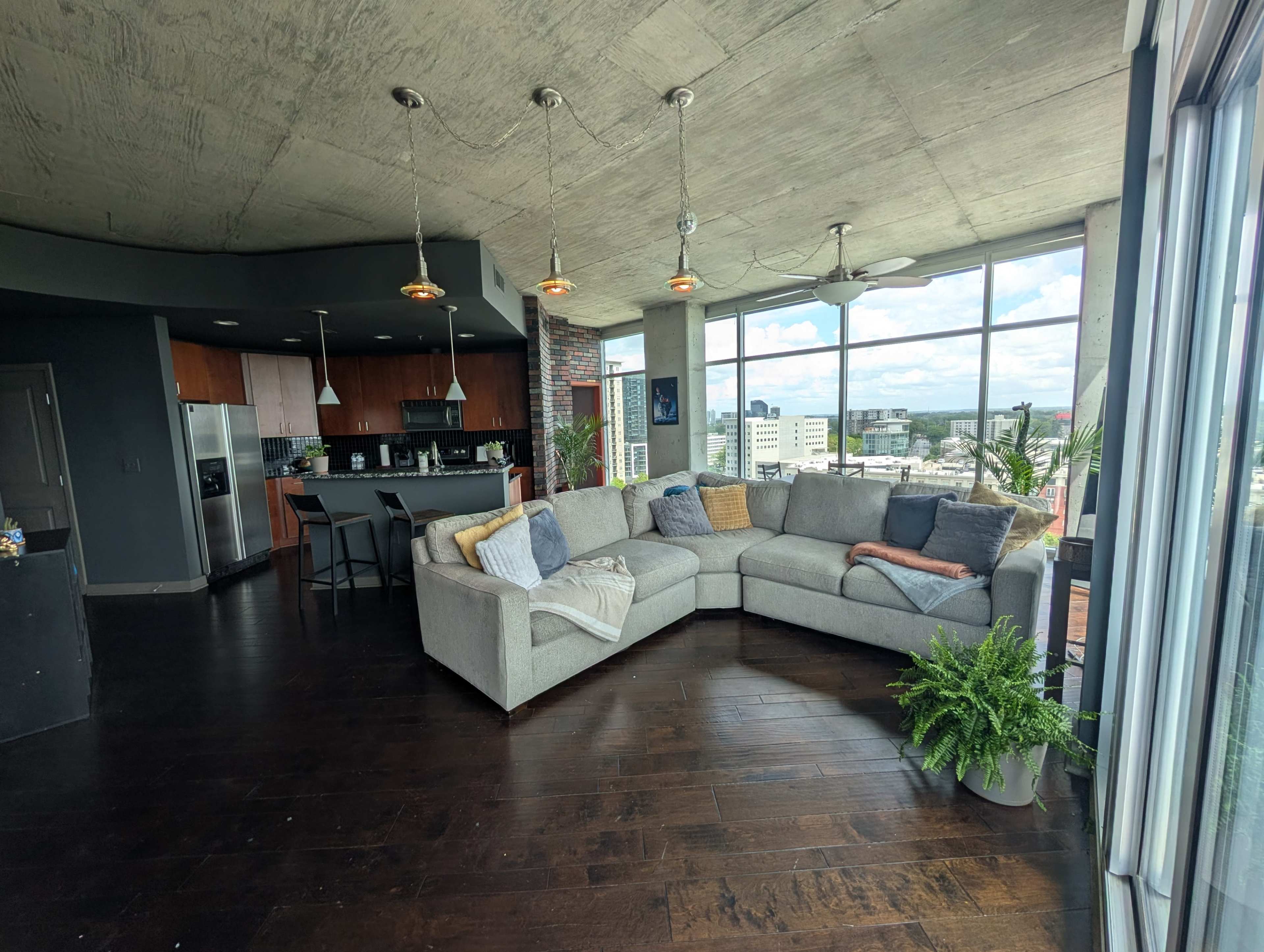 The image shows a modern living room with a large gray sectional sofa, a kitchen area in the background, and large windows that provide a view of the city.
