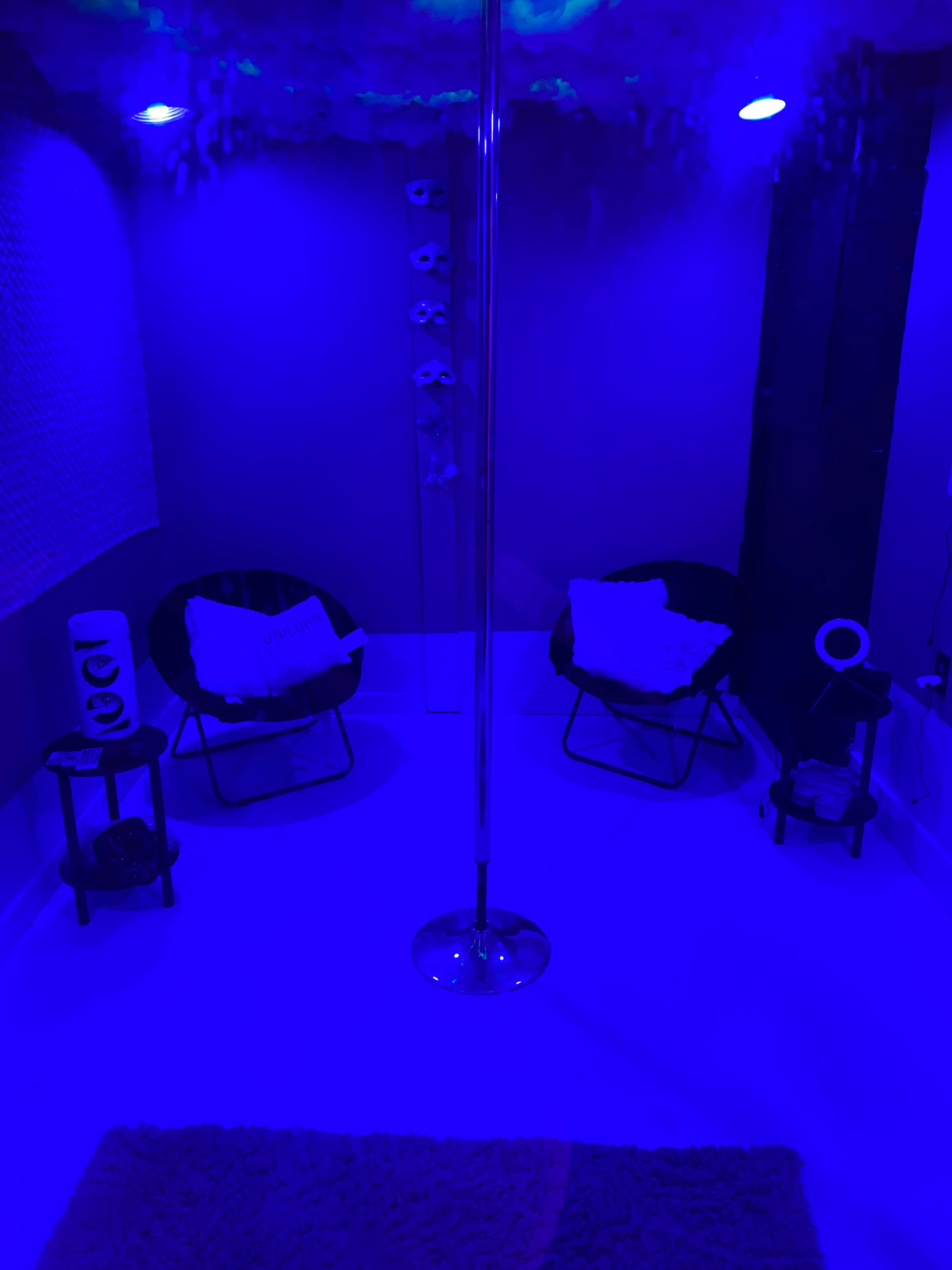 A small room features a dance pole in the center, surrounded by two black chairs, a small table, and soft lighting in shades of blue.