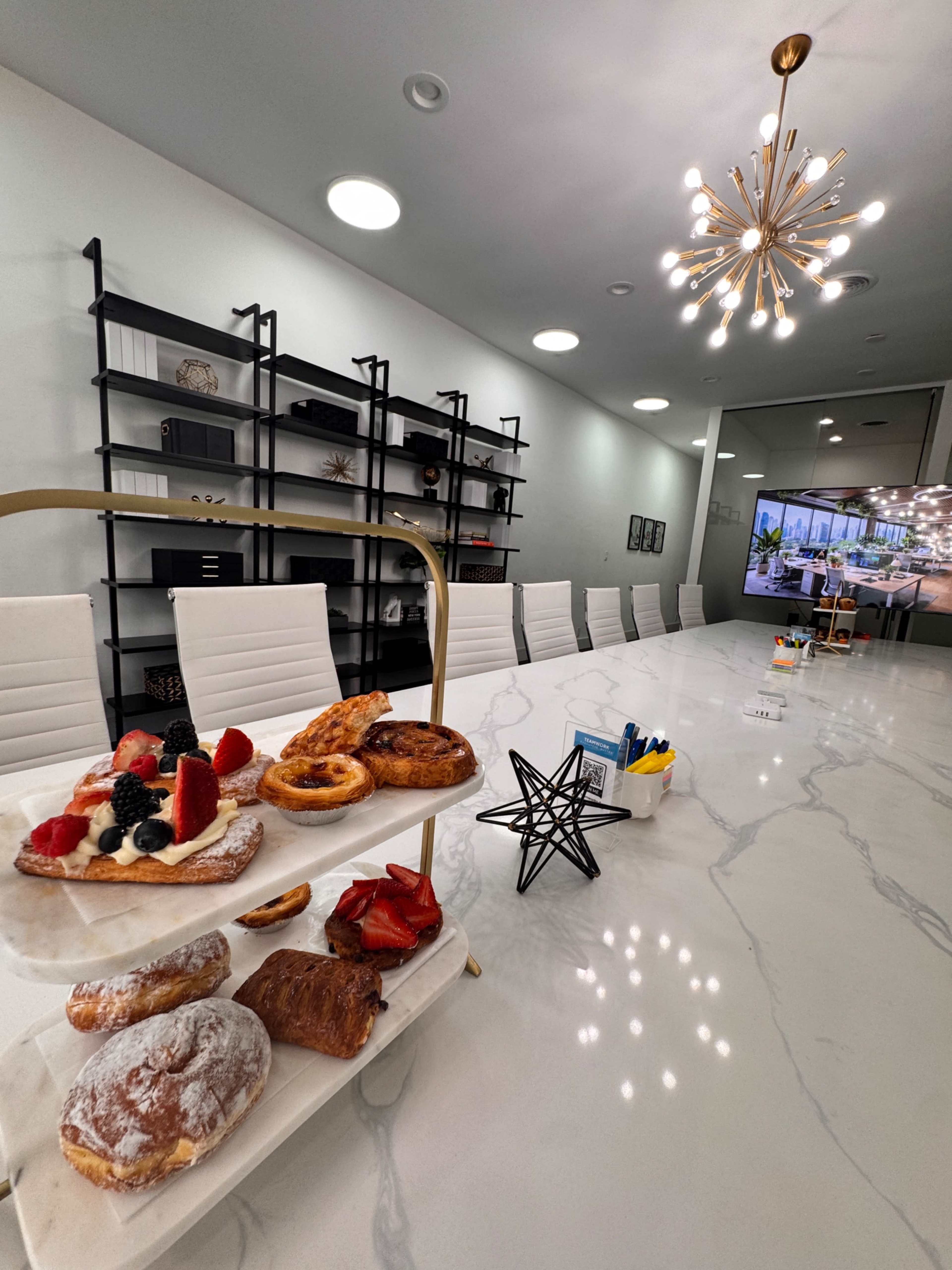 A modern conference room features a long table set with pastries and a backdrop of shelves, while a large screen displays an image of another room.
