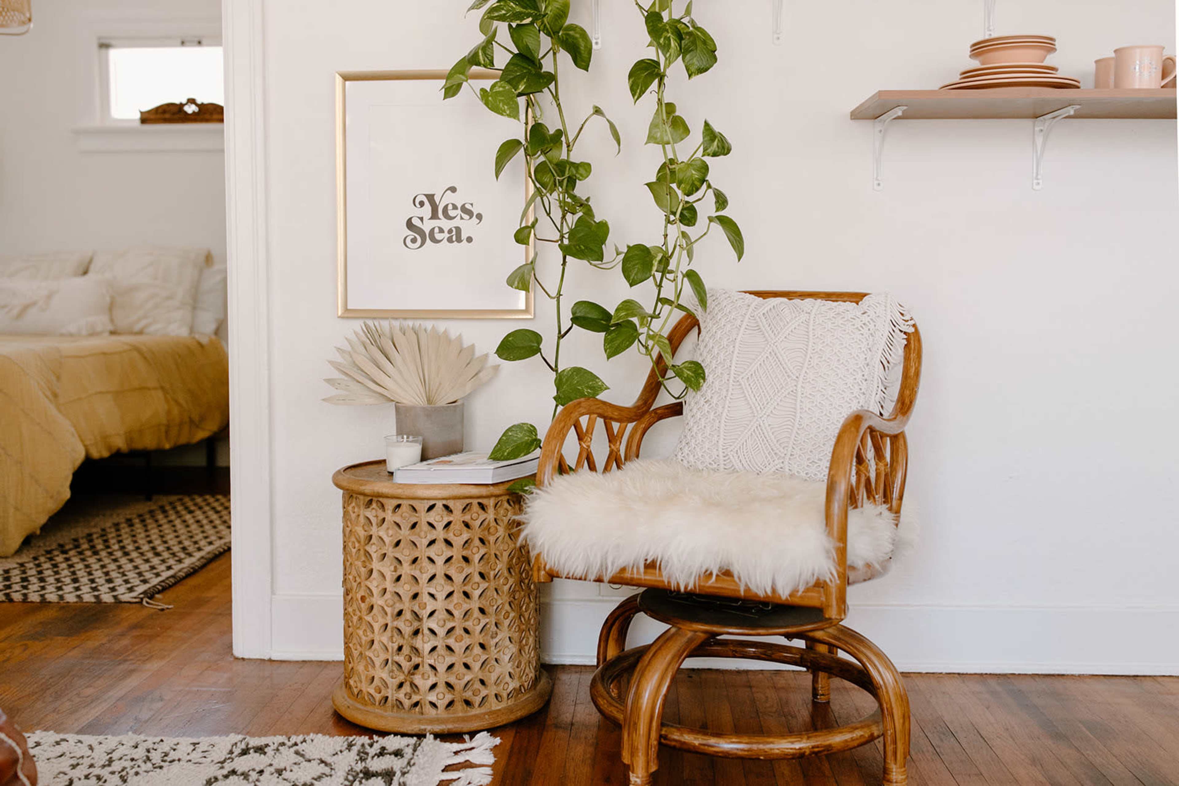A rattan chair with a fluffy white cushion is positioned next to a circular side table, adorned with a potted plant and decorative items, against a backdrop of a cozy room.