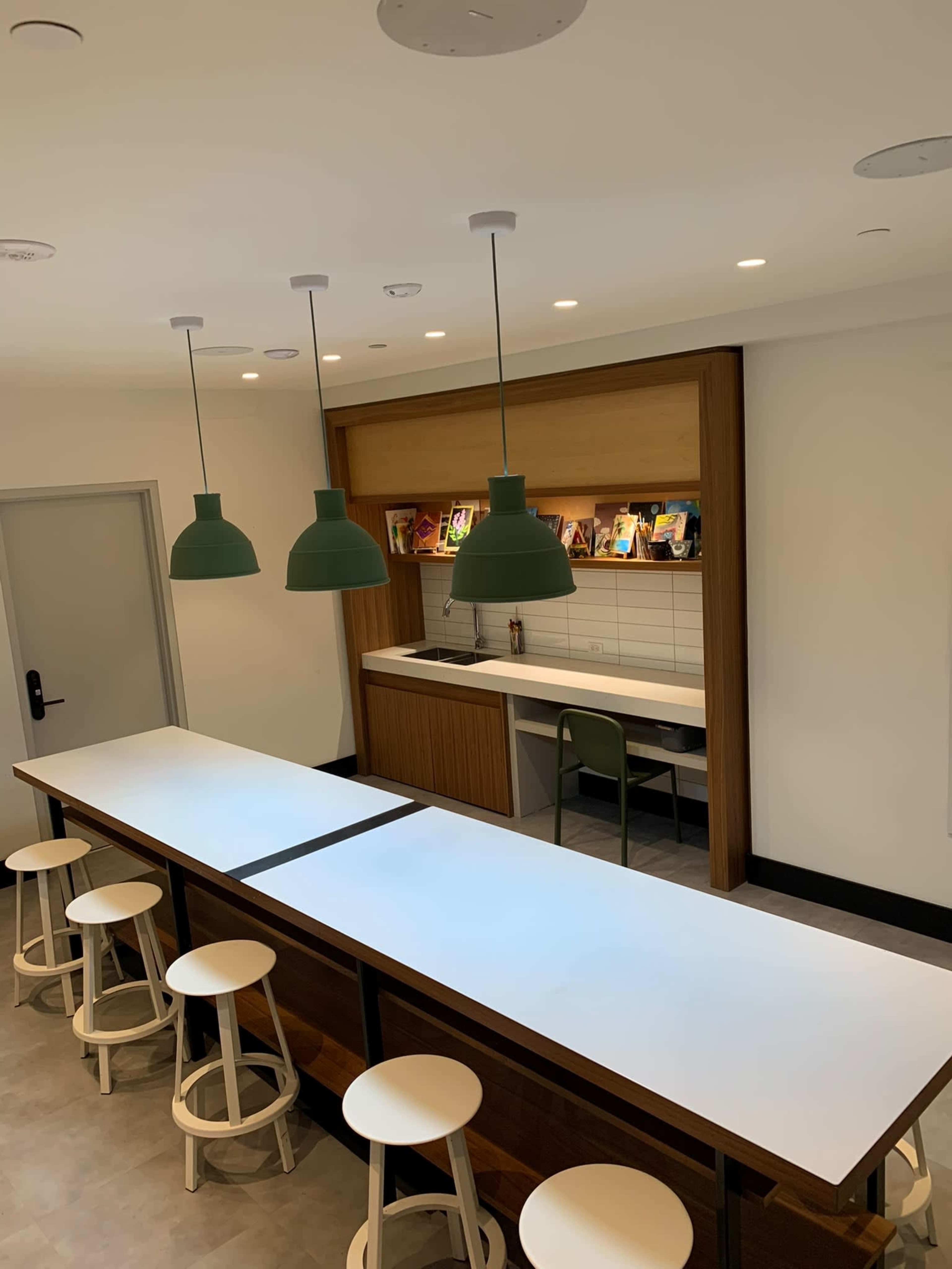 The image shows a modern kitchenette with a long, white table, green pendant lights, and white bar stools, featuring a built-in shelf with books and a sink.