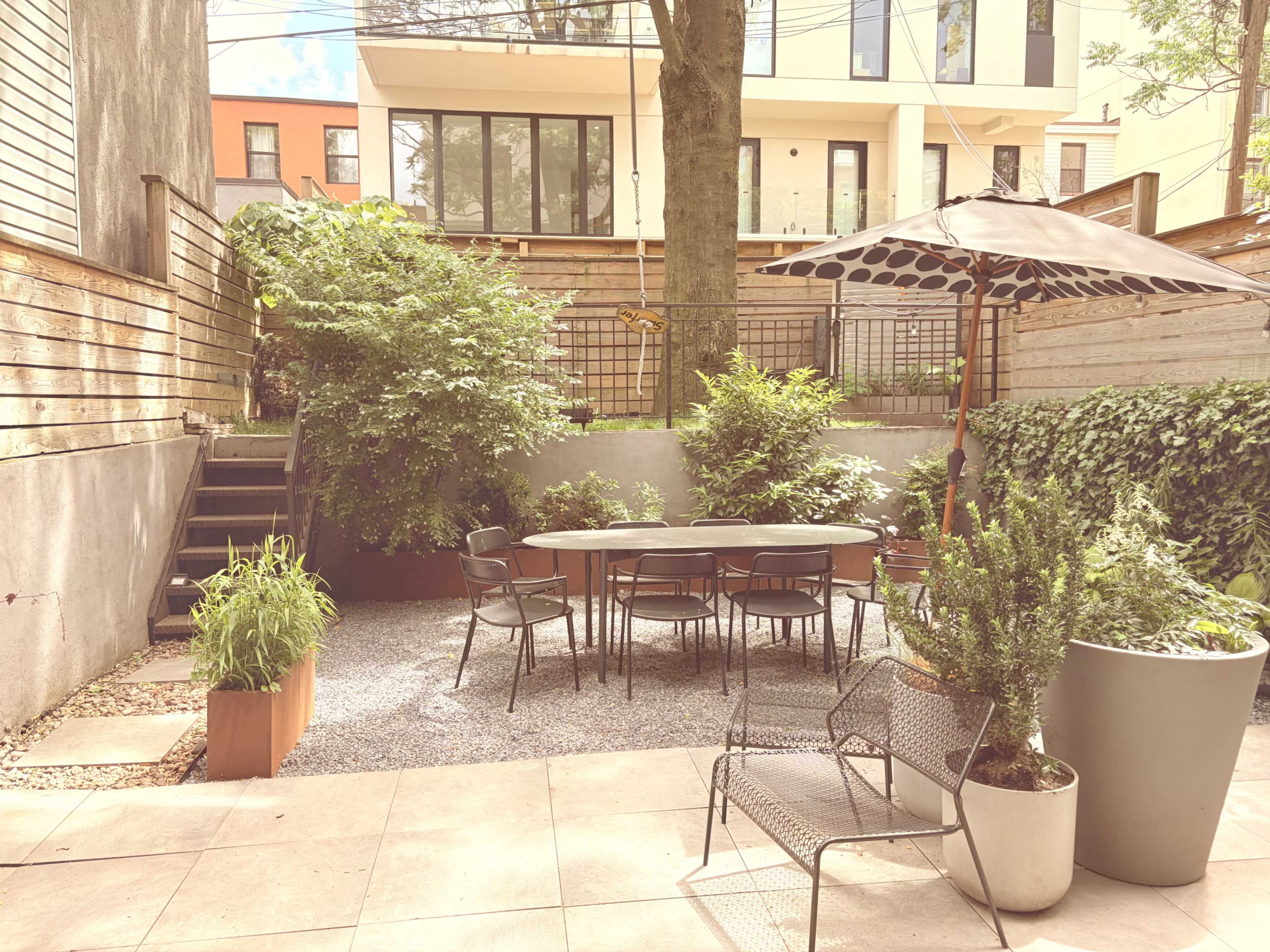 A modern garden patio features a round table surrounded by black chairs, with potted plants and an umbrella amidst lush greenery and gravel.