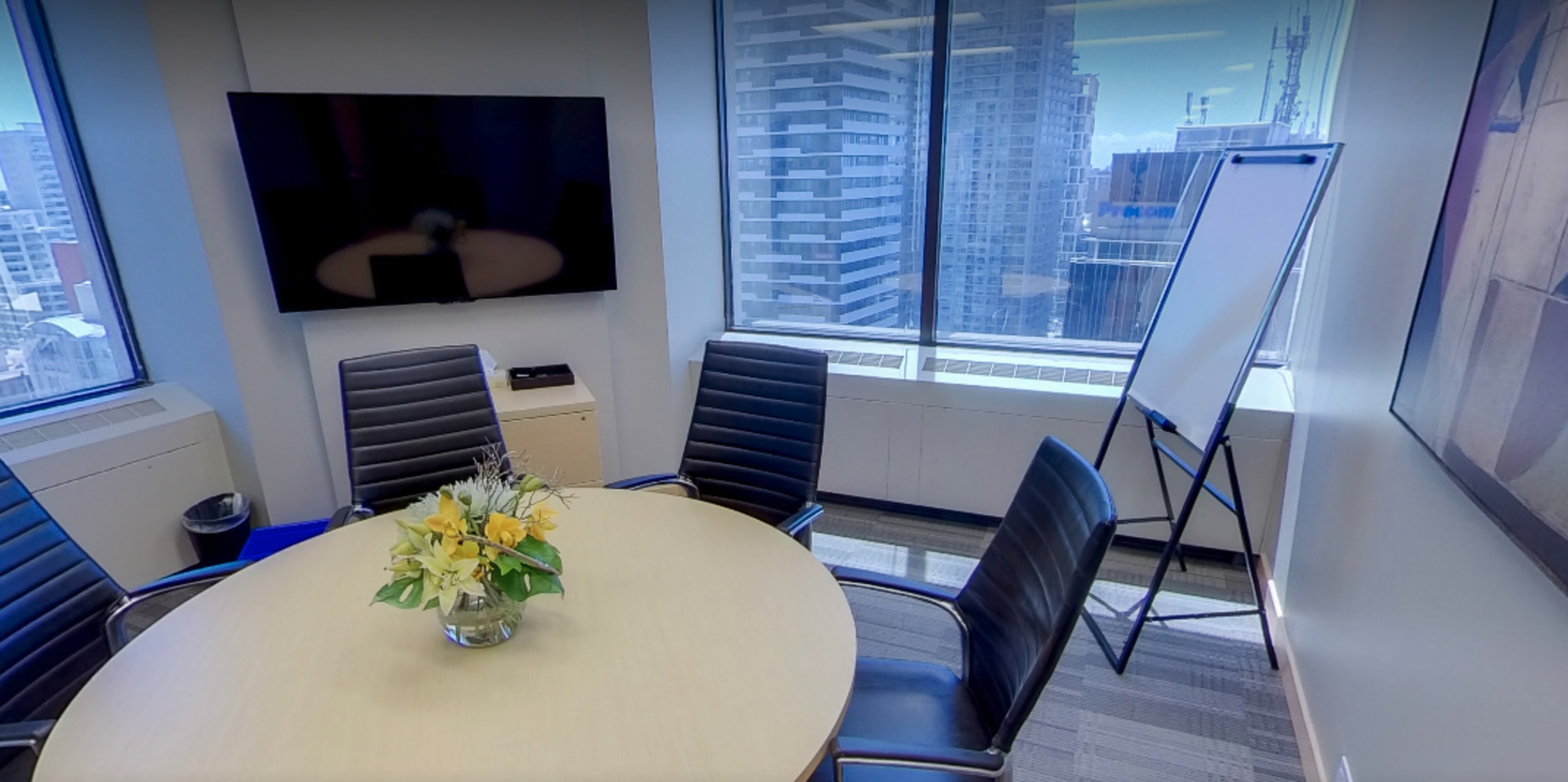 A conference room features a round table with a floral centerpiece, four black chairs, a whiteboard stand, and large windows showcasing a cityscape.