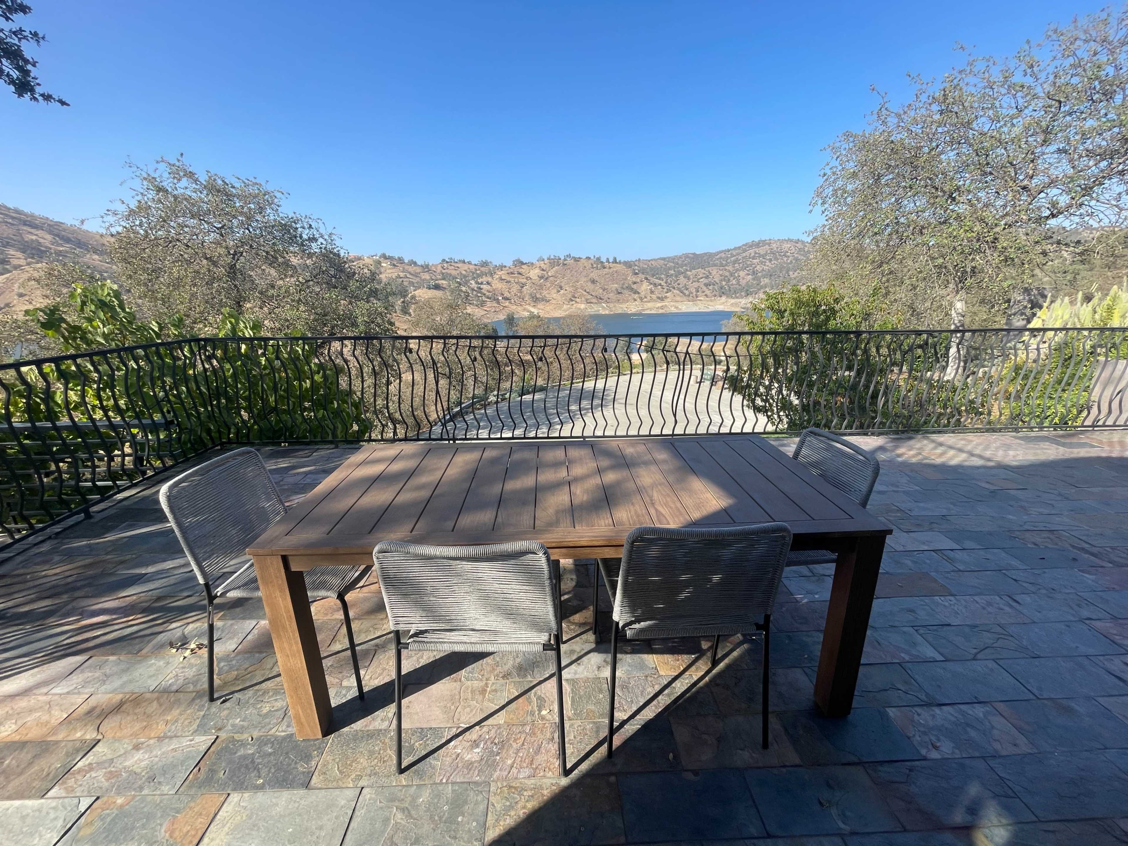 A wooden table with four chairs is set on a stone-paved patio, overlooking a lake and hillside under a clear blue sky.