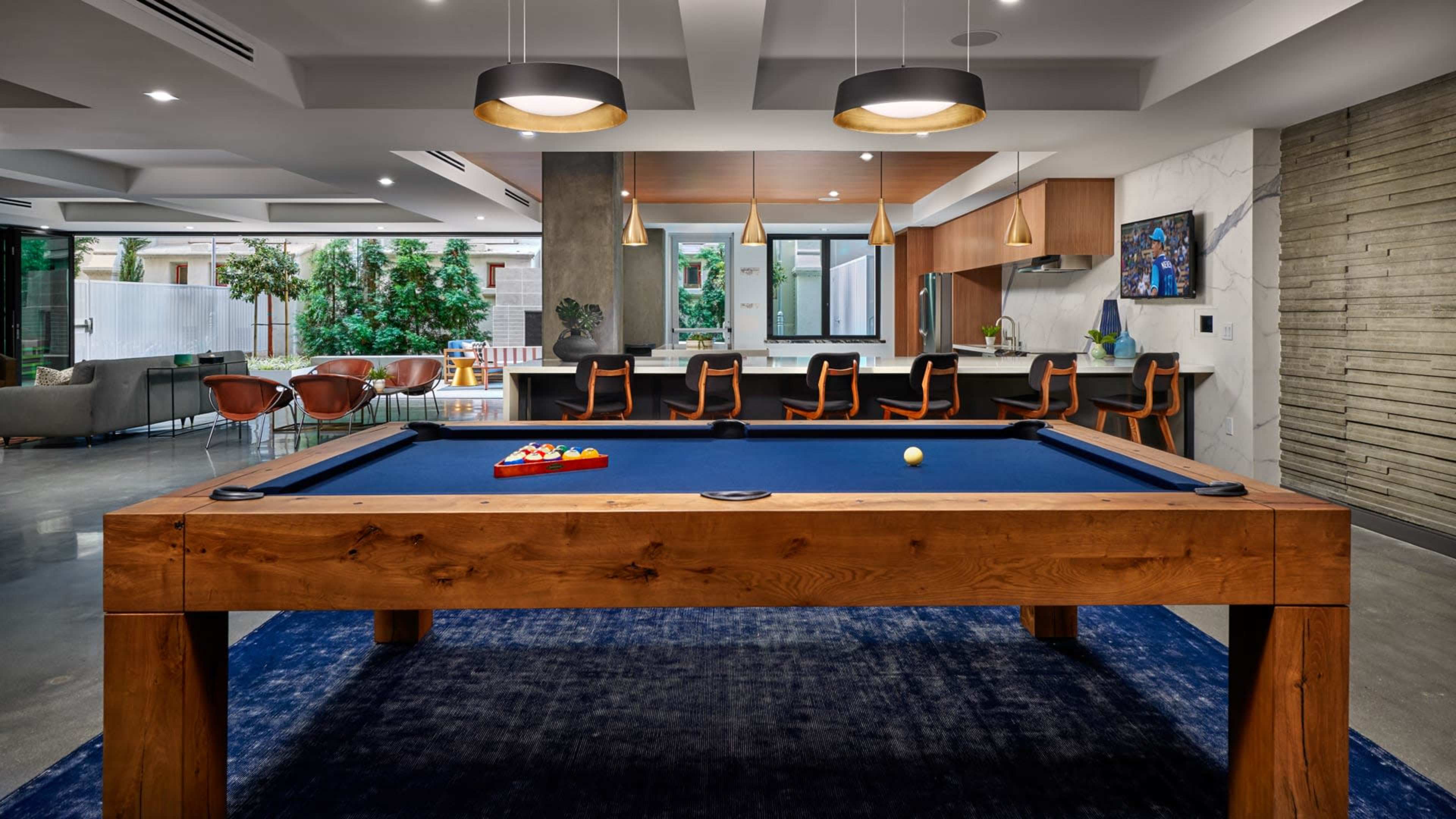 A modern game room featuring a large wooden pool table surrounded by a blue rug, with a kitchen and dining area visible in the background.