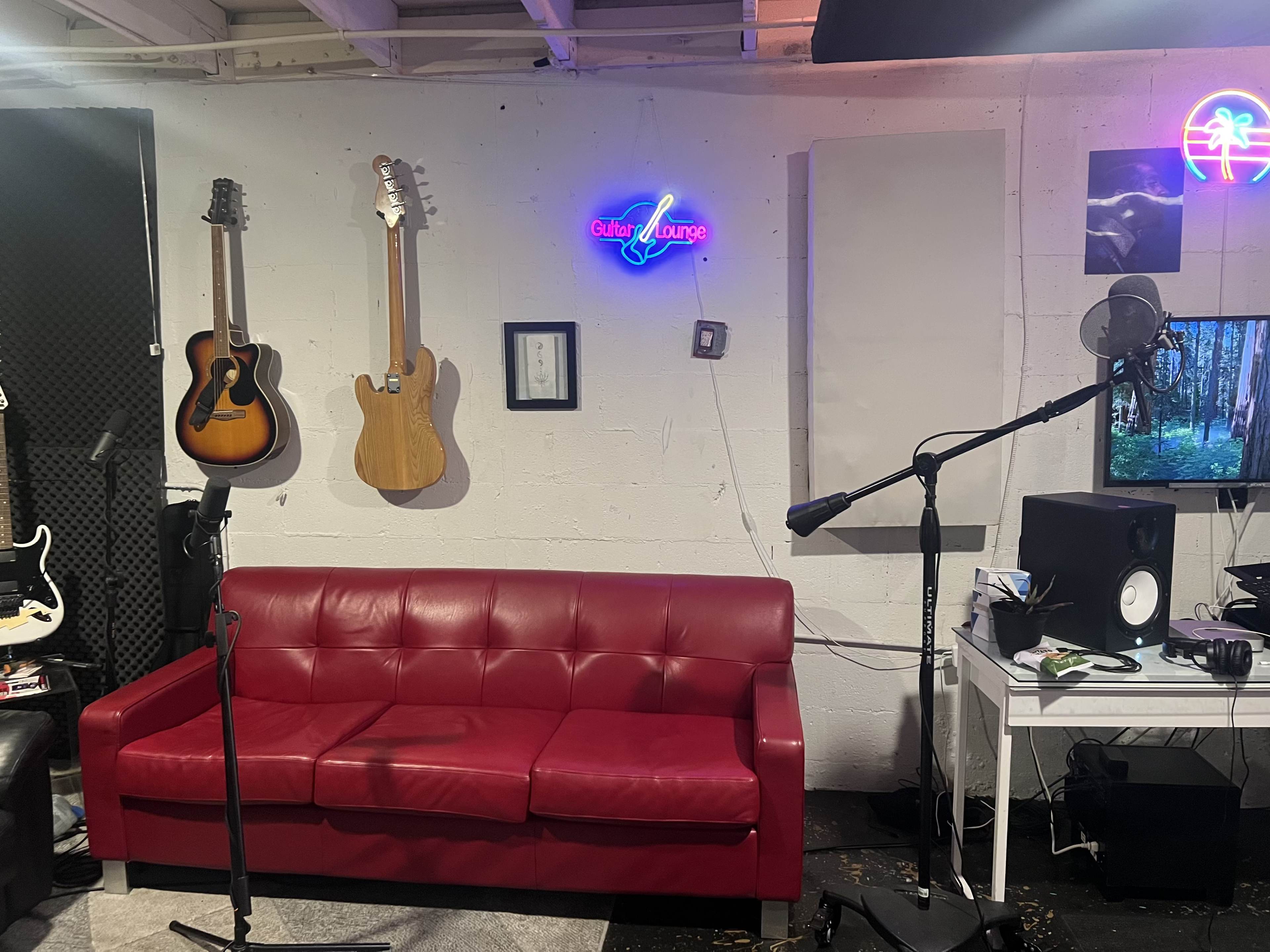 The image shows a music room with a red leather couch, several guitars hanging on the wall, a microphone on a stand, and a computer setup with speakers and headphones.