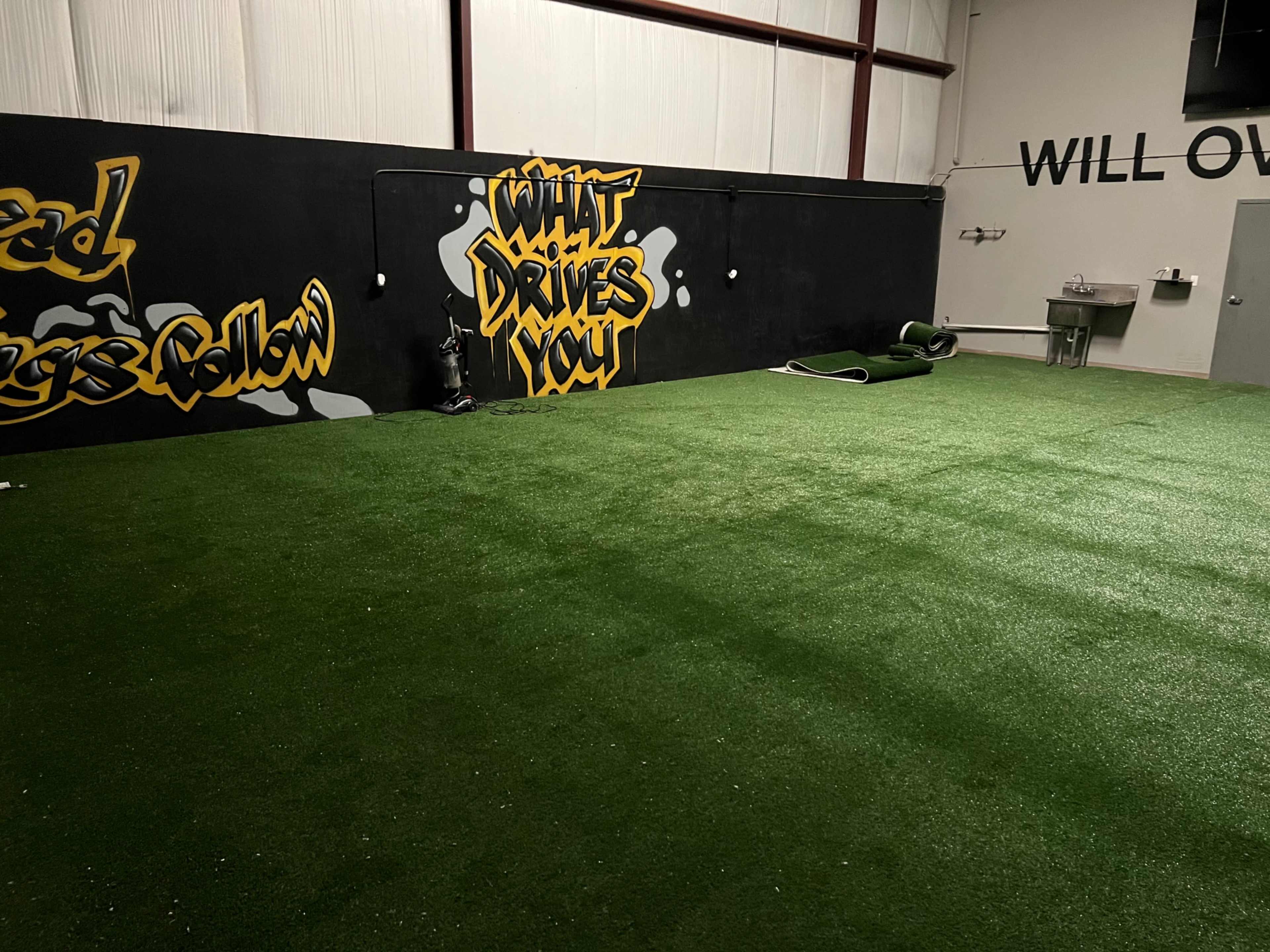 A spacious indoor area with artificial turf, black walls featuring colorful graffiti, and a sink area in the corner.