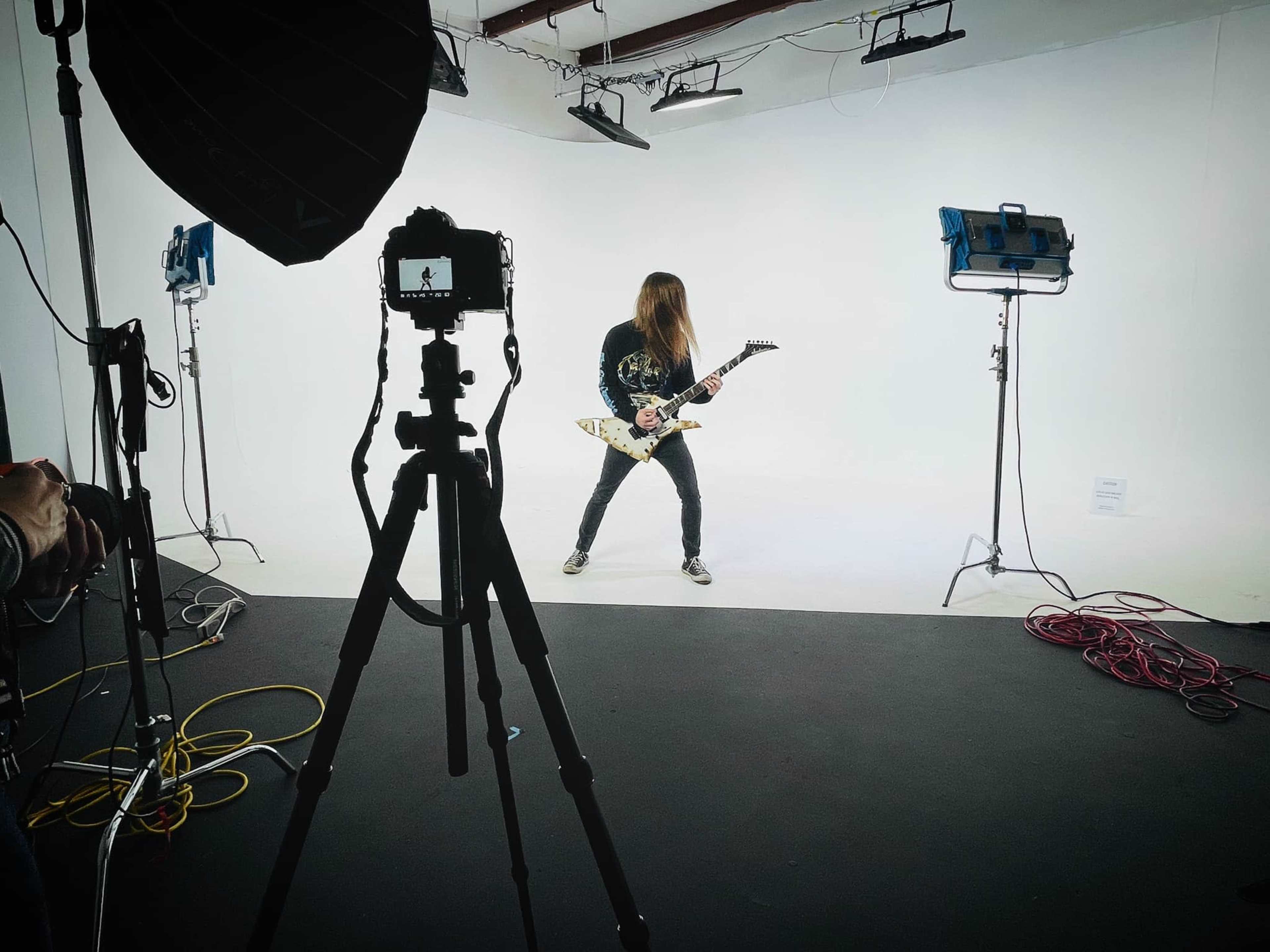 A guitarist stands in a studio space with a white backdrop, illuminated by several lights, while being photographed from a distance.
