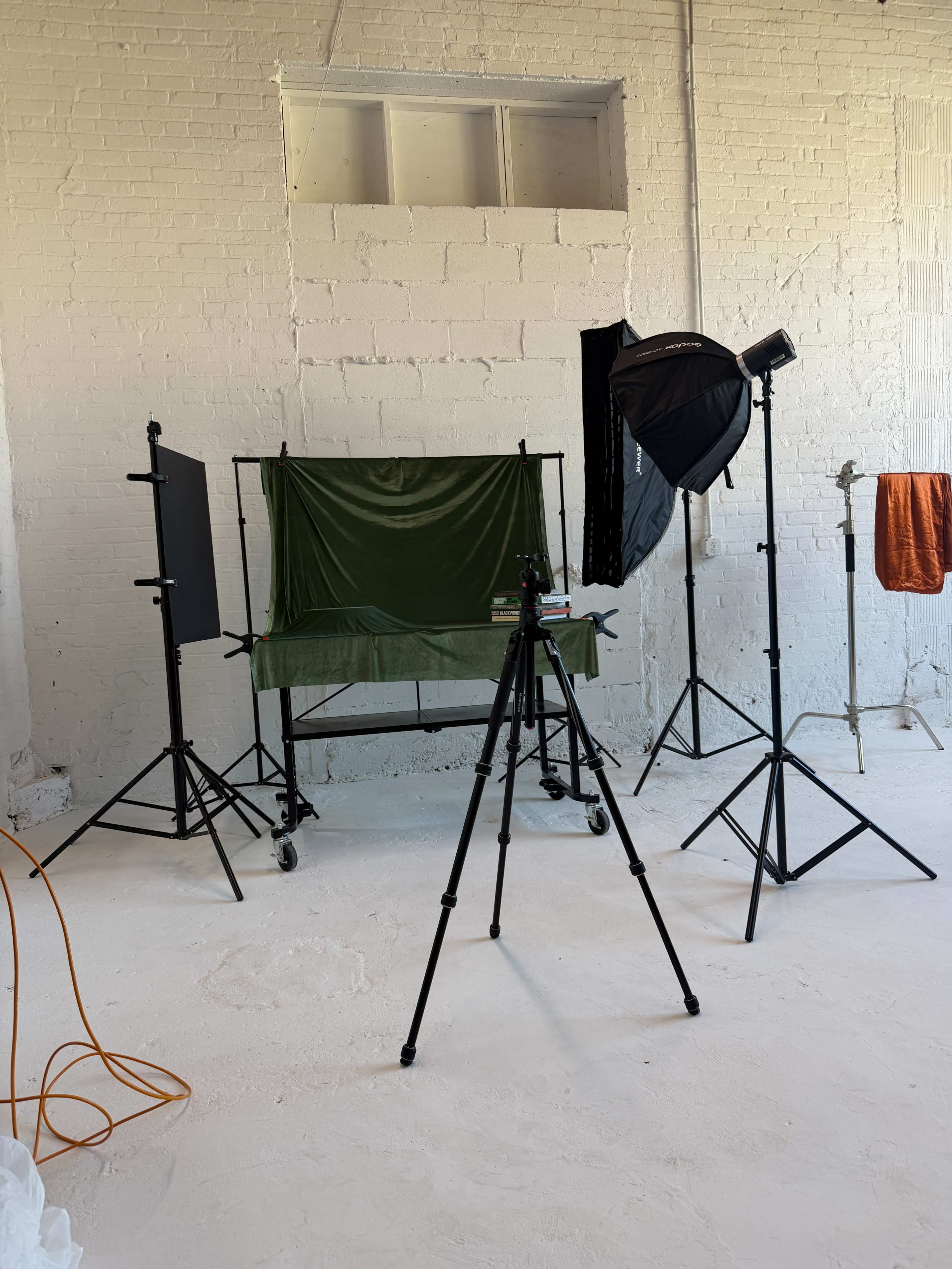 The image shows a photography setup in a studio, featuring a green backdrop on a stand, a tripod with a camera, and various lighting equipment.
