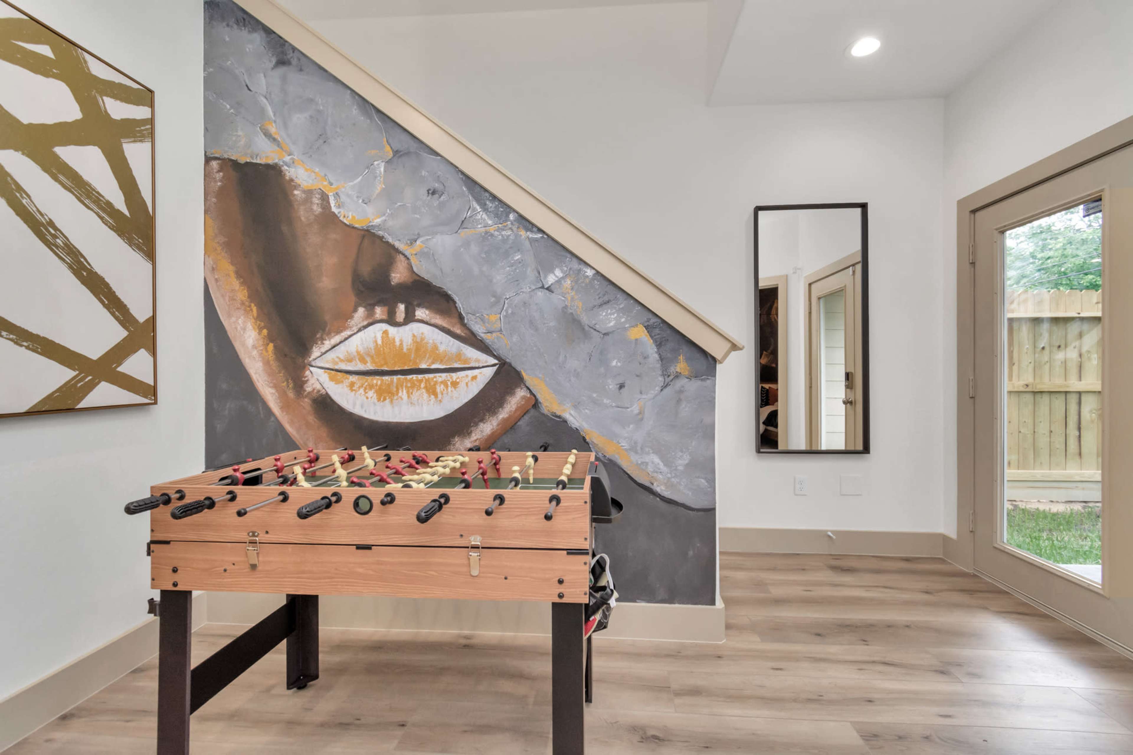 A foosball table sits near a staircase, accompanied by a large wall mural and a mirror in a well-lit room.