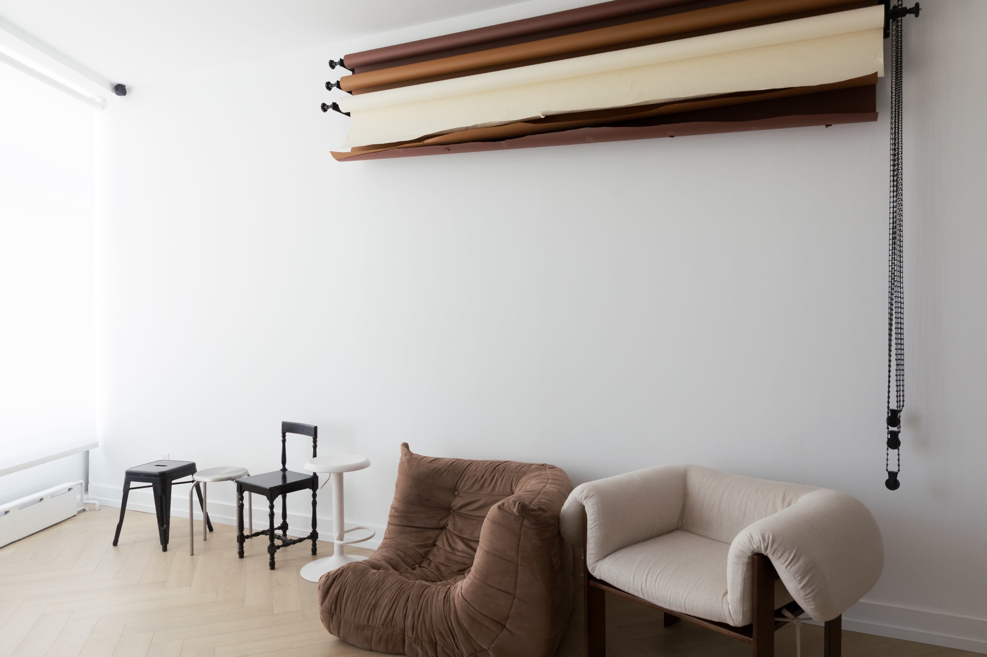 The image shows a minimalist interior with two distinct chairs, a small round table, and a roll of backdrop paper mounted on the wall.