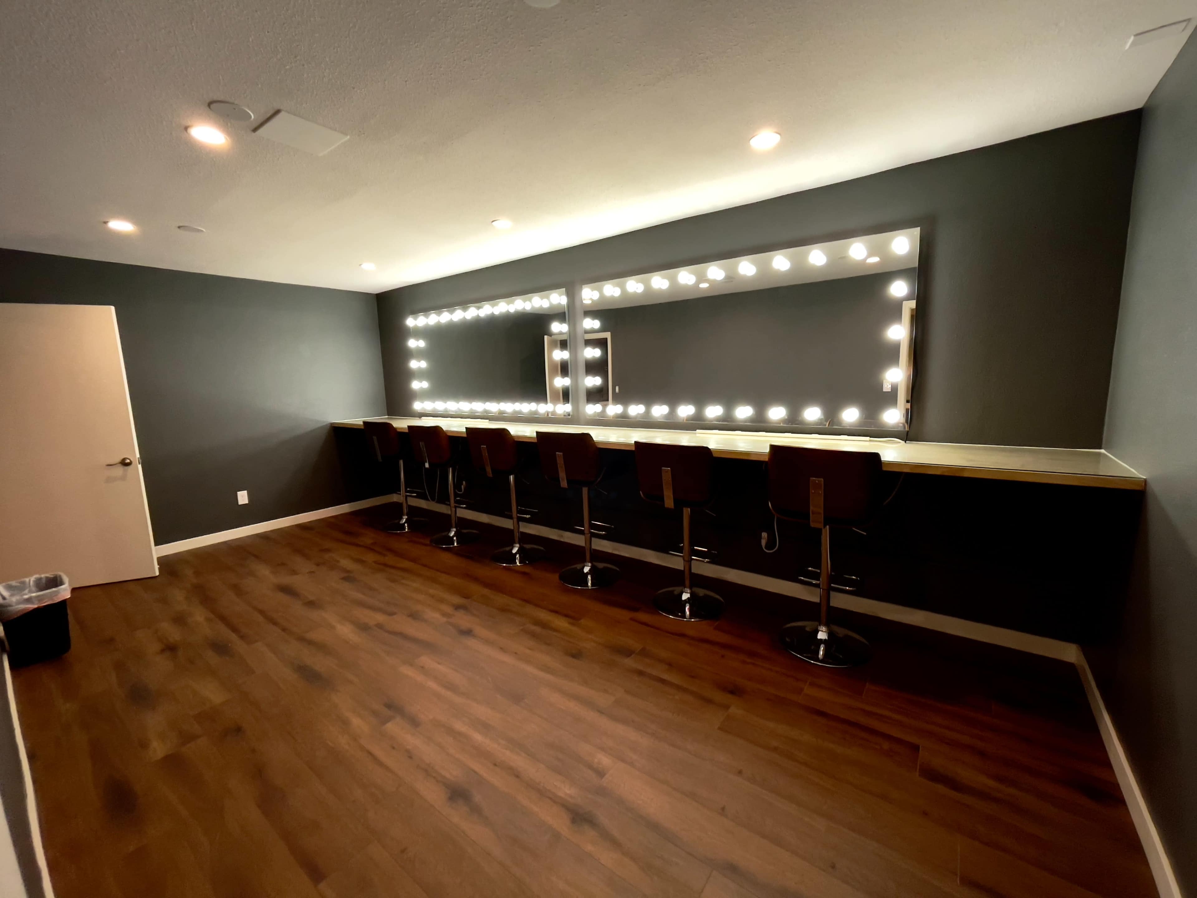 A well-lit dressing room with a mirrored vanity and several barstools positioned along a wooden counter.
