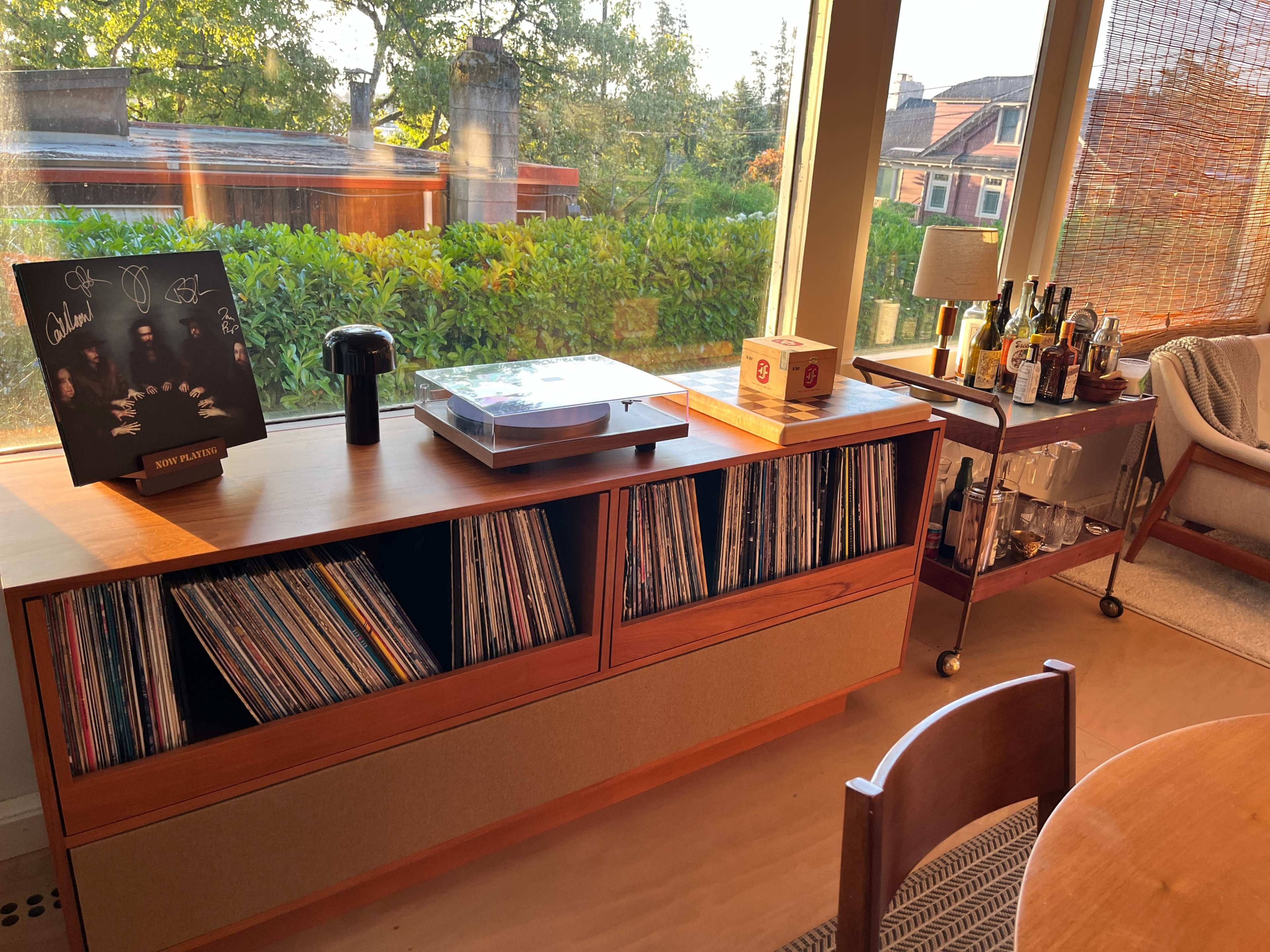 The image shows a wooden record storage unit filled with vinyl records, accompanied by a turntable, a black speaker, and a cart displaying various bottles, all set against a backdrop of a large window with greenery outside.
