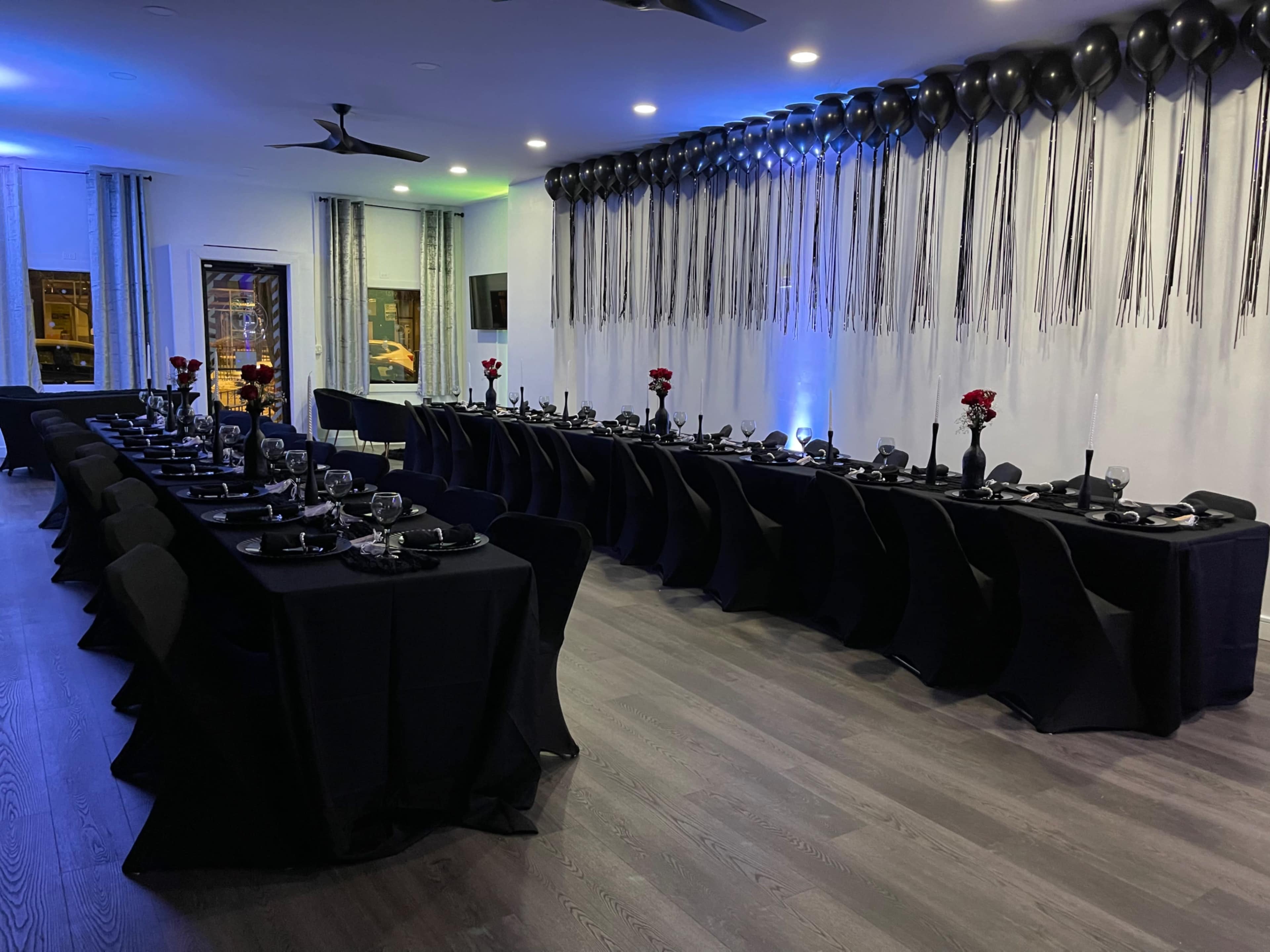 A long banquet table is set with black tablecloths, elegant dinnerware, and red roses, surrounded by chairs, in a dimly lit room decorated with black balloons and silver streamers.