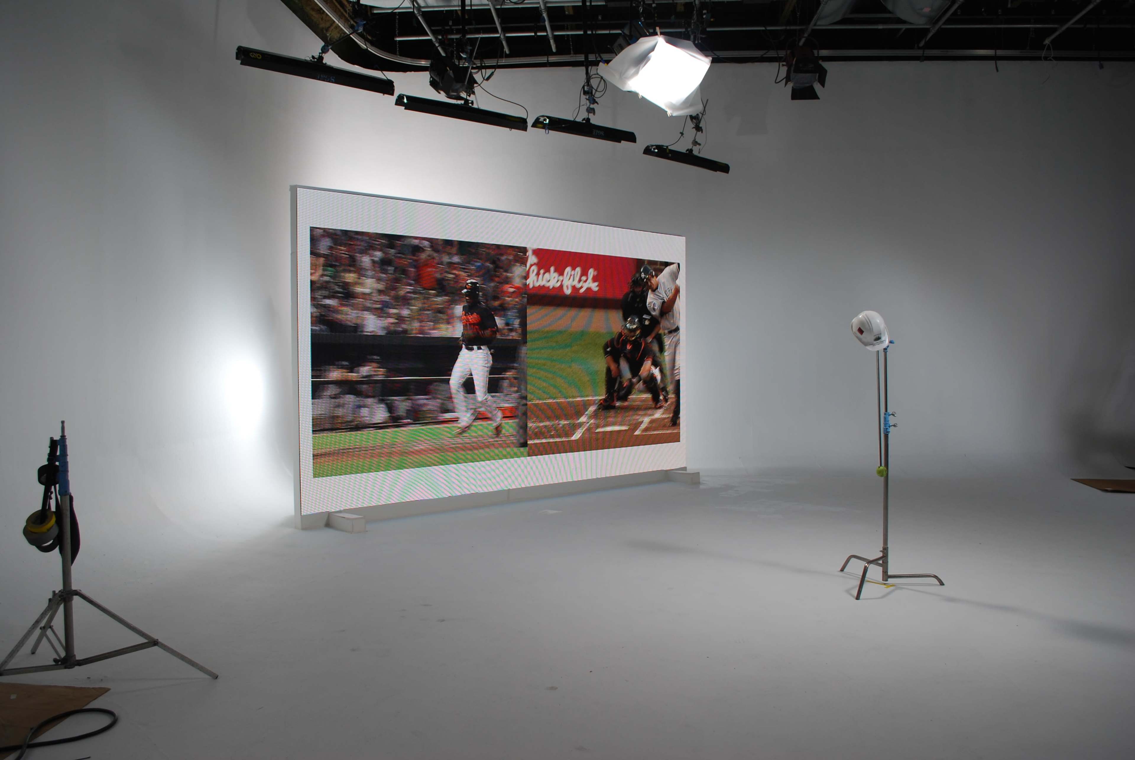 A large digital display shows two images side by side, one of a sports player in action on a field and the other capturing a player at bat in a baseball game, set in a studio with bright lighting and equipment.